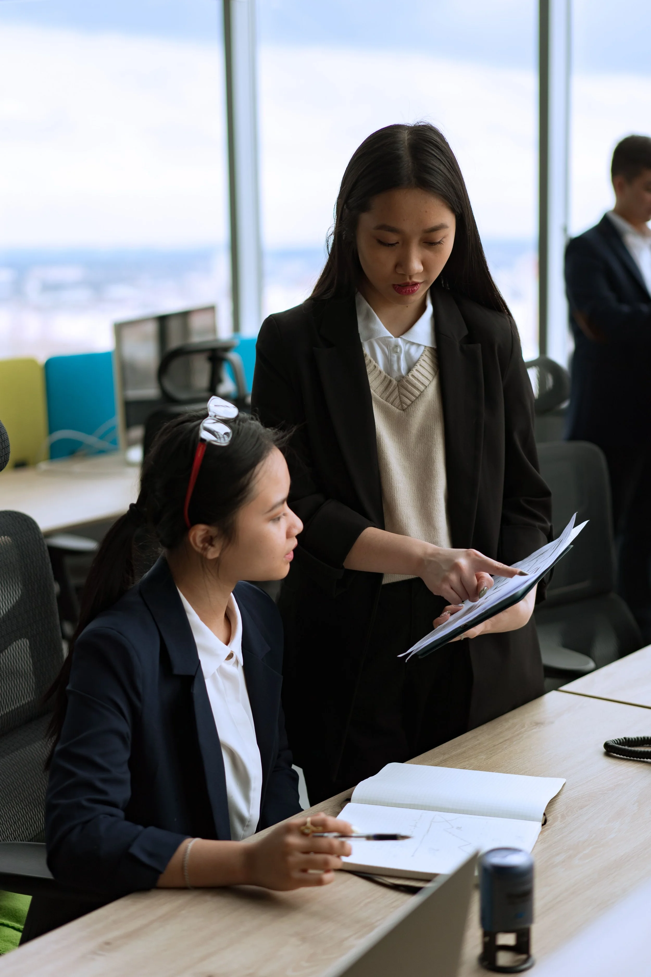 Businesswomen in a modern office engaged in a discussion, with one woman standing and holding documents while talking to another woman seated at a conference table, with notebooks and office supplies on the table, and large windows showing a cityscape in the background.