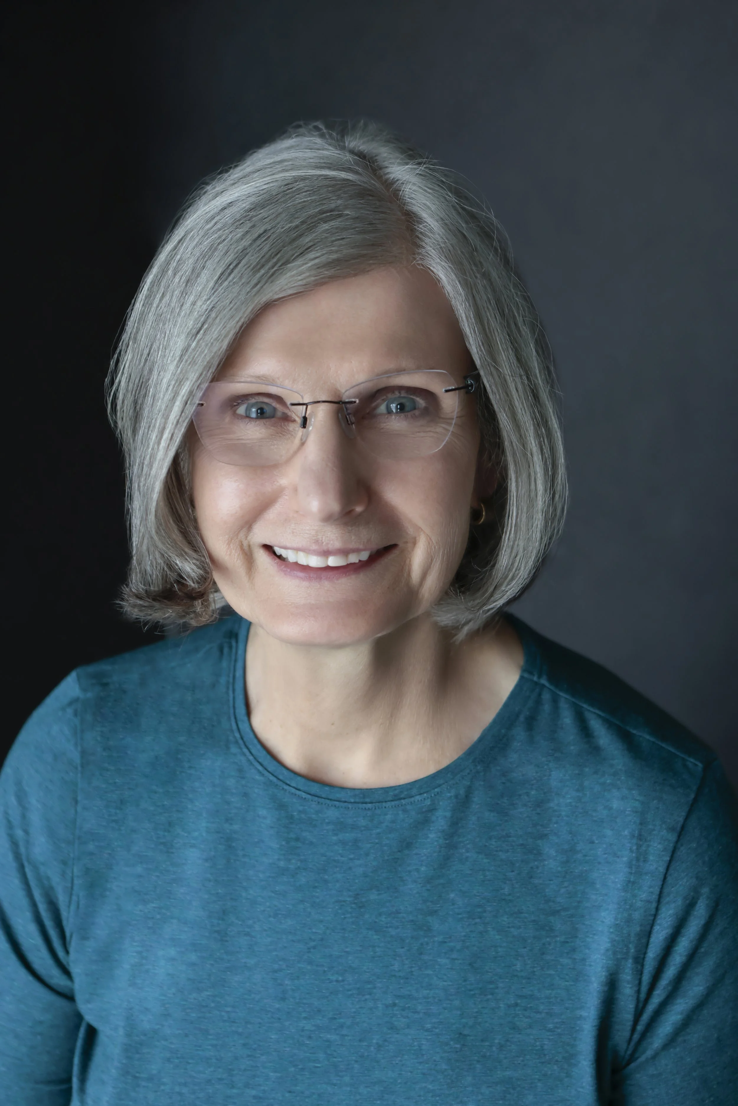 Smiling older woman with gray hair and glasses wearing a teal shirt