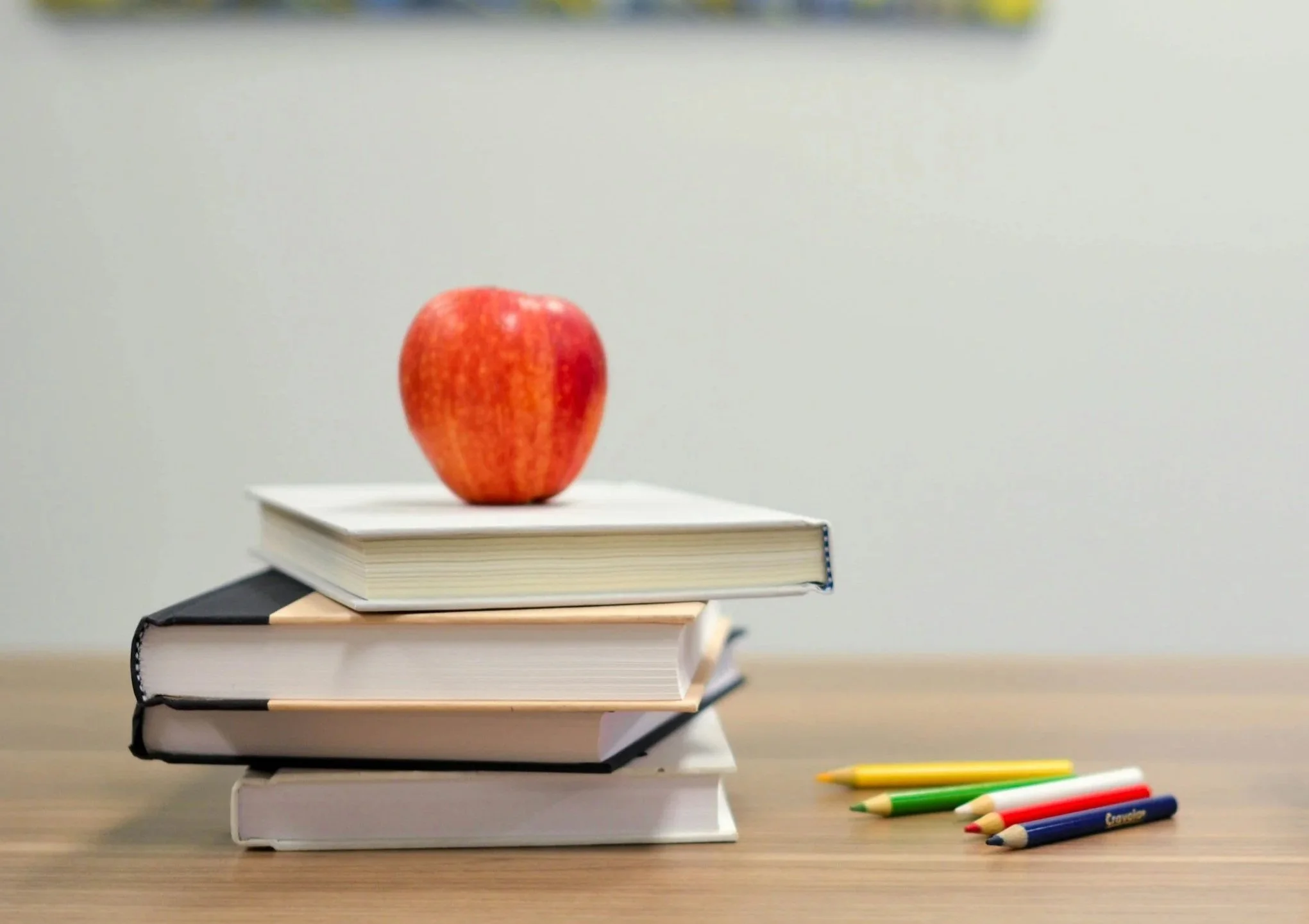 A red apple placed on top of a stack of four closed books on a wooden surface, with colored pencils nearby.