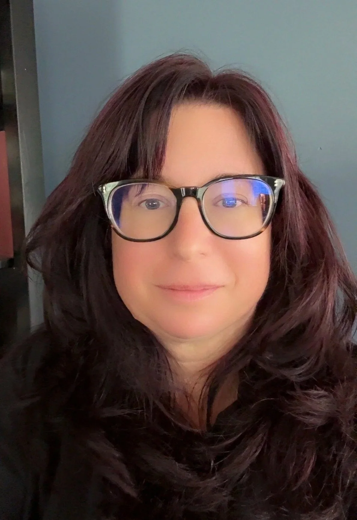 A woman with long, dark auburn hair and glasses takes a selfie indoors, background in shades of green and gray.