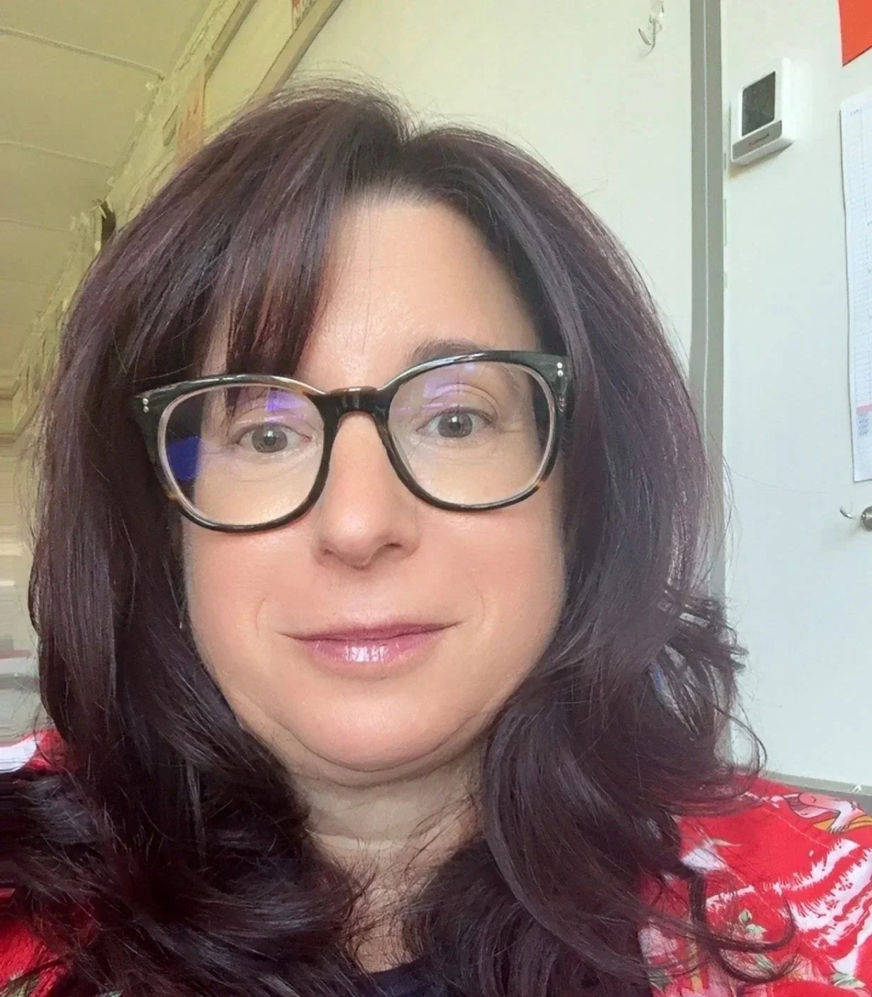 Close-up photo of a woman with wavy, auburn hair and glasses, smiling softly, indoors with a white wall, thermostat, and a paper on the wall in the background.