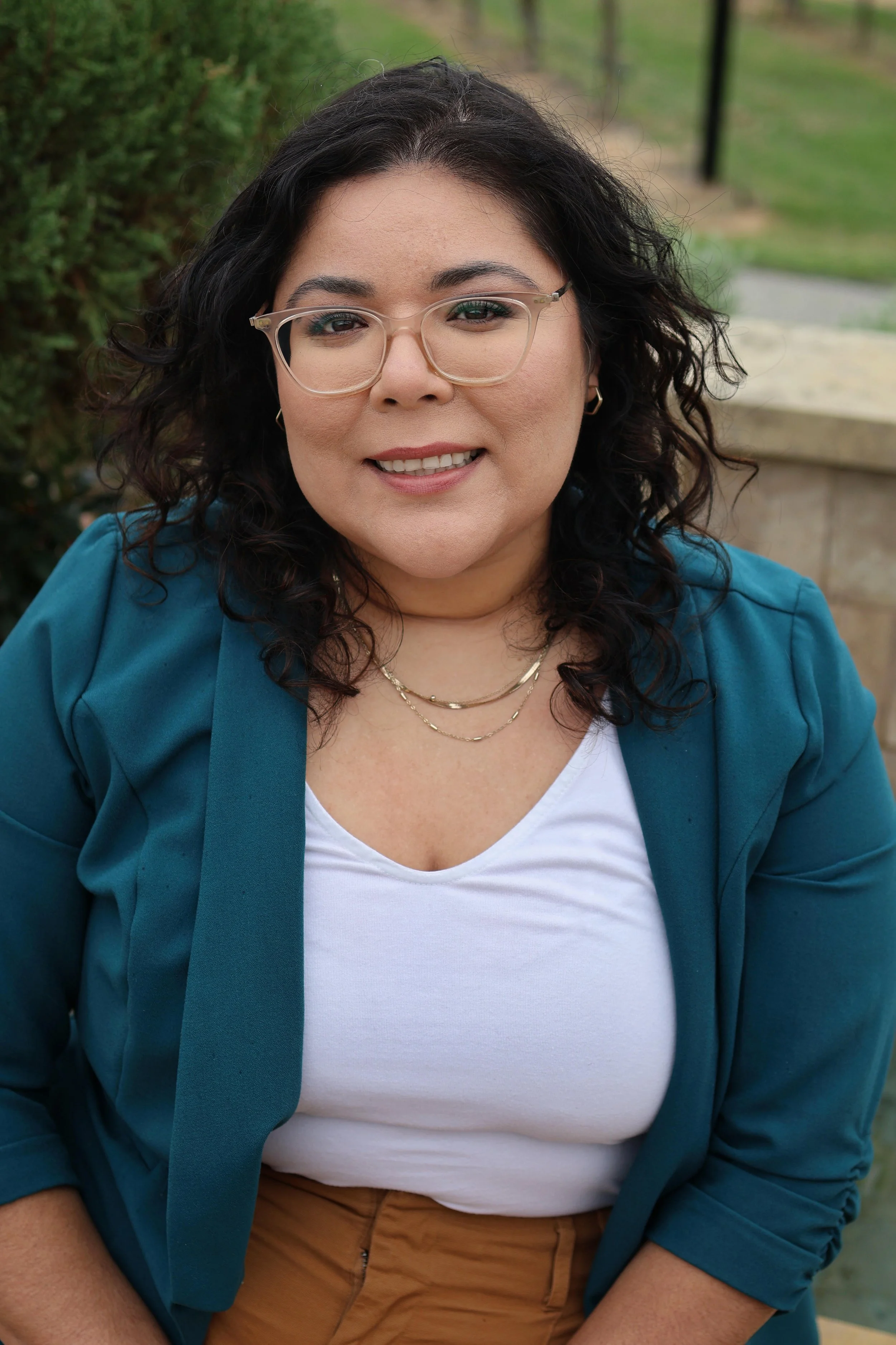A woman with curly dark hair and glasses smiling outdoors, wearing a blue blazer, white top, and tan pants.