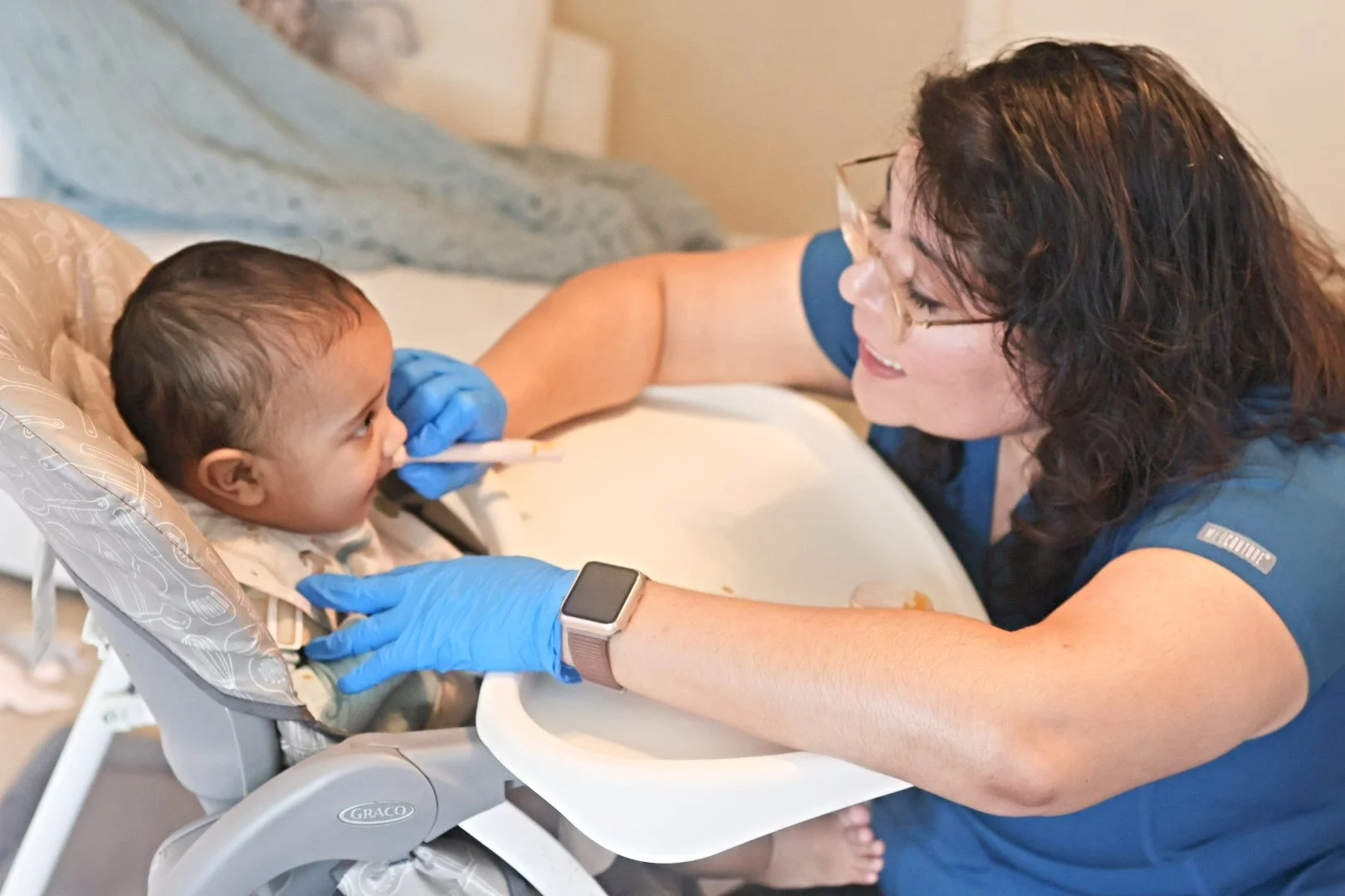 A healthcare worker in blue scrubs and gloves gives a nasal swab to a young child sitting in a high chair, smiling during a health checkup.