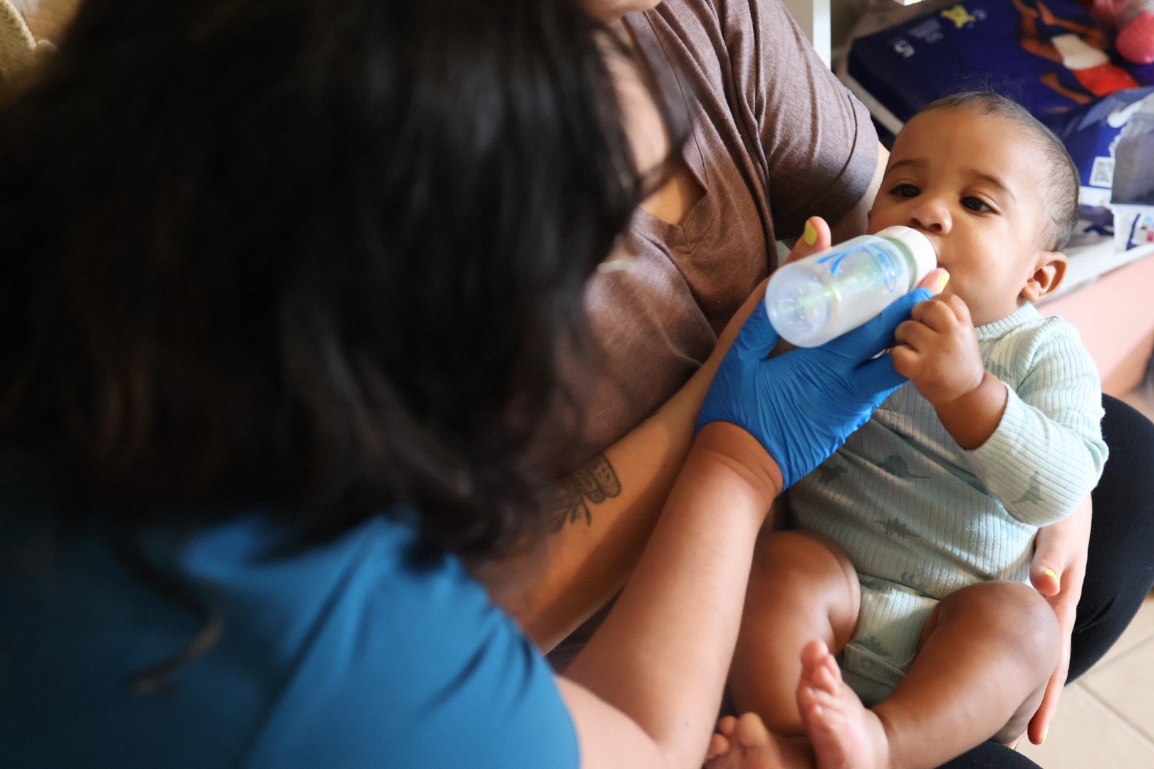 A woman is feeding a baby with a bottle while another adult assists, in a cozy indoor setting.