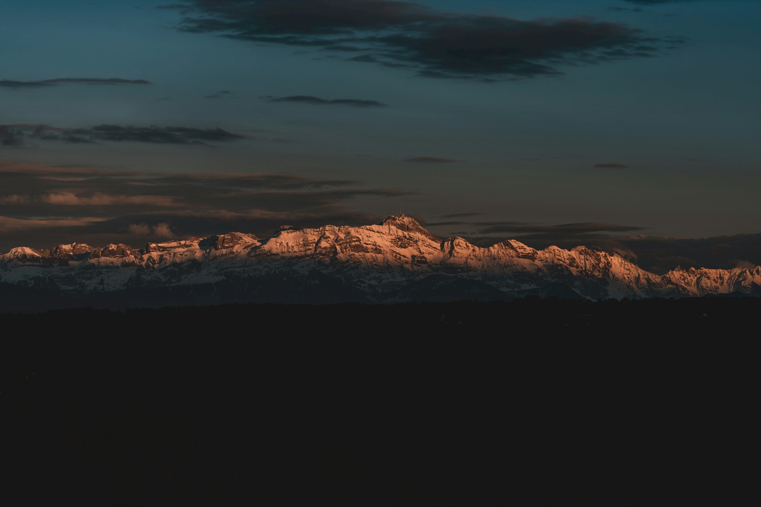 Snow-capped mountain range under a dark cloudy sky at dusk.