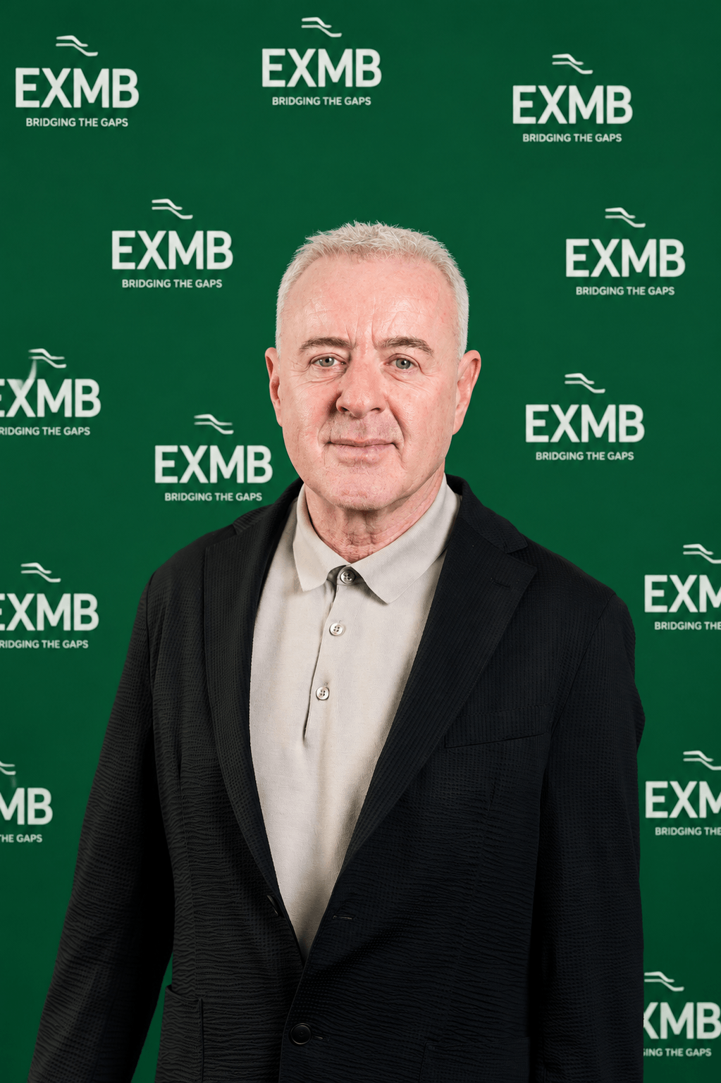 Steve Bridge standing in front of a green backdrop with the EXMB logo and the slogan "Bridging the Gaps" printed multiple times. He has short grey hair and is wearing a light-coloured shirt with a dark blazer.