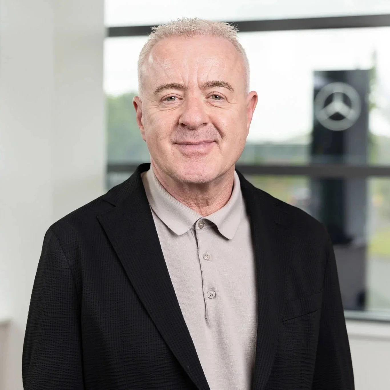 Steve Bridge  standing in front of large windows in an office environment, with the Mercedes-Benz logo in the background. He is wearing a beige collared shirt and a black blazer, smiling softly at the camera.