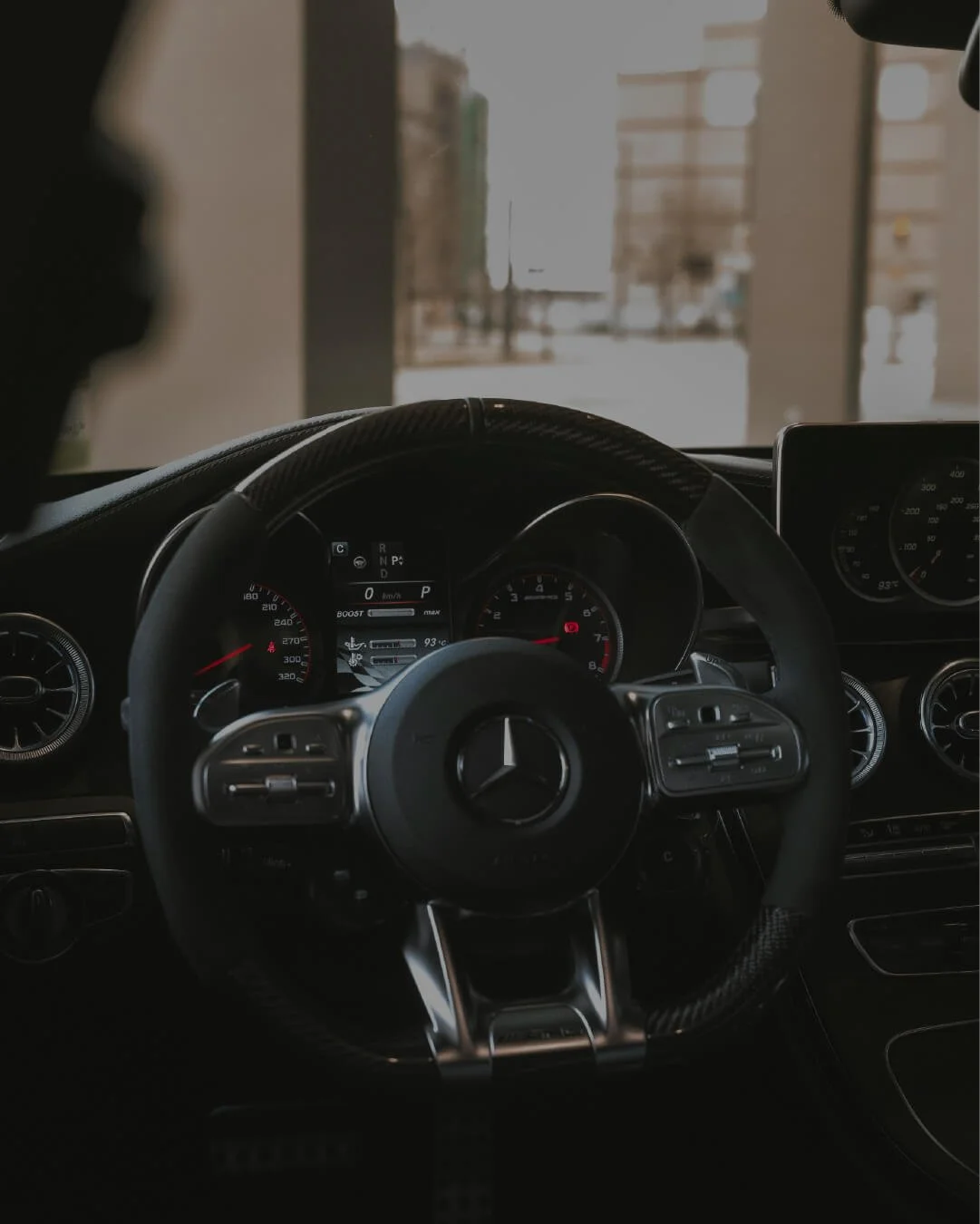 Close-up of a Mercedes-Benz car steering wheel and dashboard, showing speedometer, tachometer, and infotainment screen, with a view outside through the windshield.