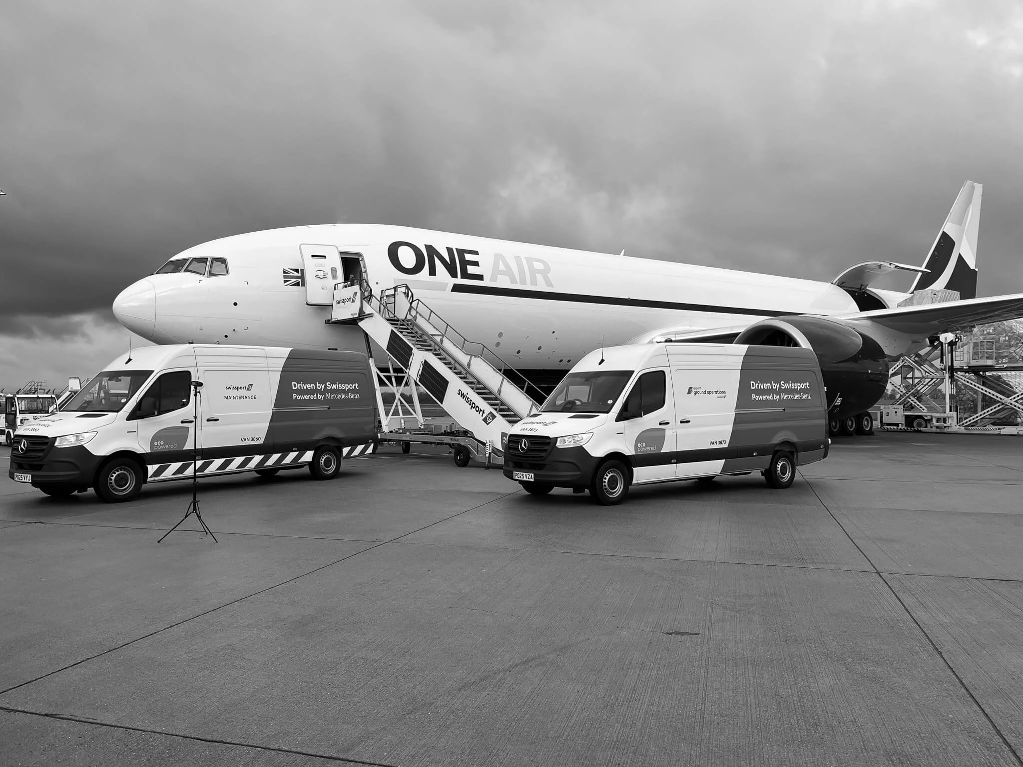 A black and white photograph of an airplane on the tarmac with Mercedes-Benz vehicles under a cloudy sky.