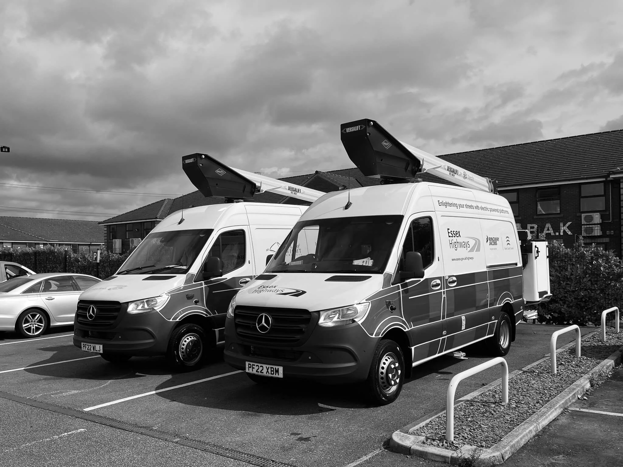 Two white electric utility Mercedes-Benz vans with roof-mounted charging stations parked in a parking lot, residential buildings in the background, cloudy sky overhead.