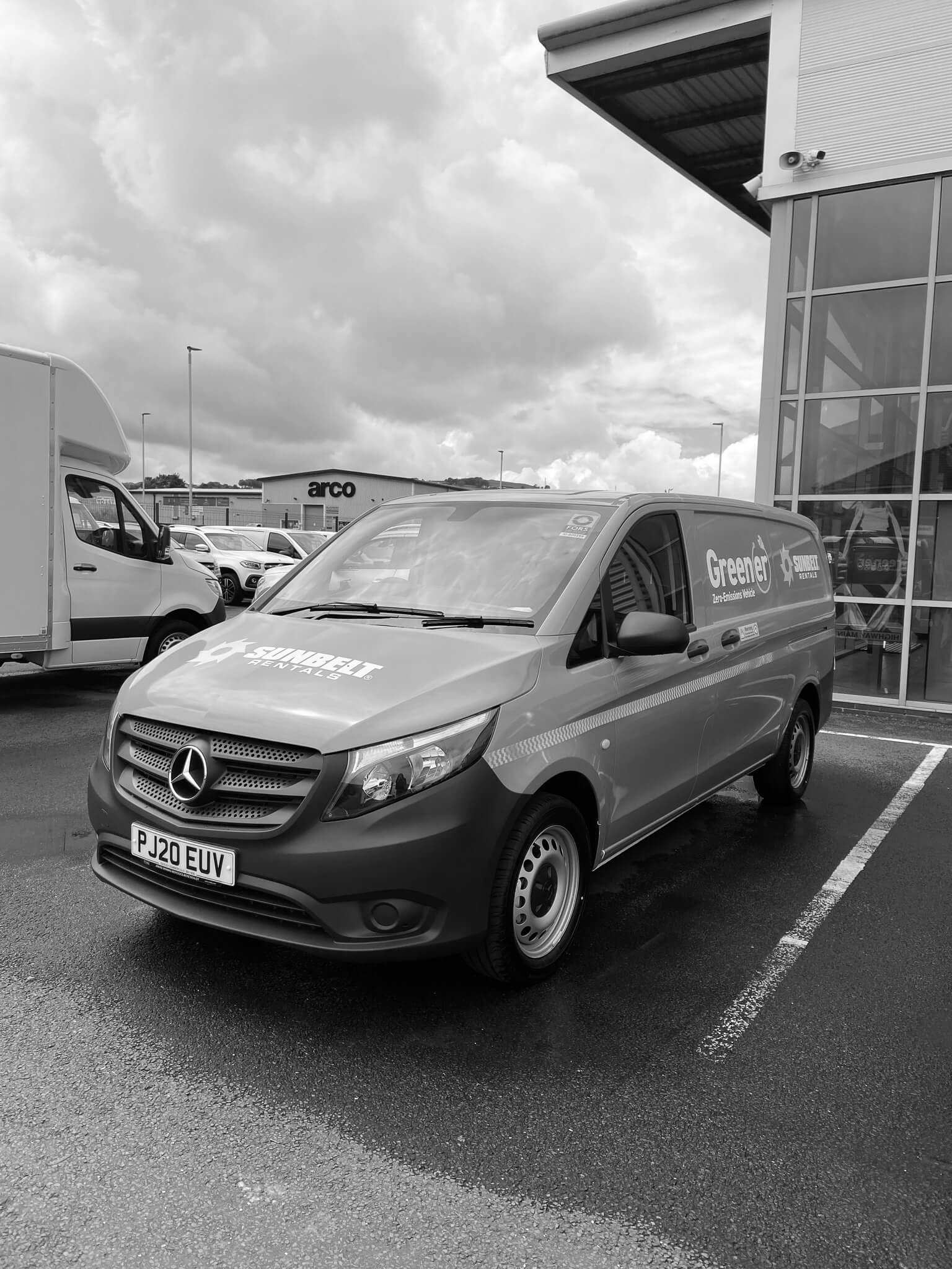 Black and white photo of a parking lot outside a building with a large glass window. Several cars are parked, including a Mercedes-Benz van in the foreground. The building has a sign that reads 'arco' in the background.