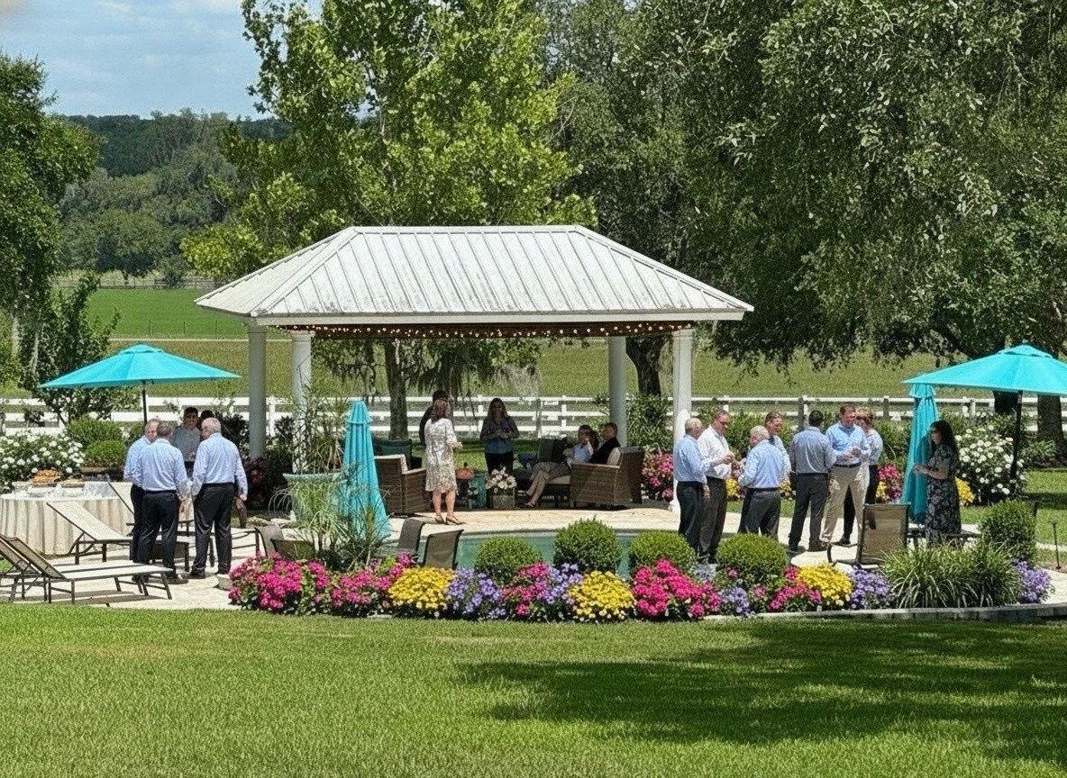 A group corporate event people gathered around a gazebo in a lush green outdoor setting with flowers and trees, some chatting and others sitting.
