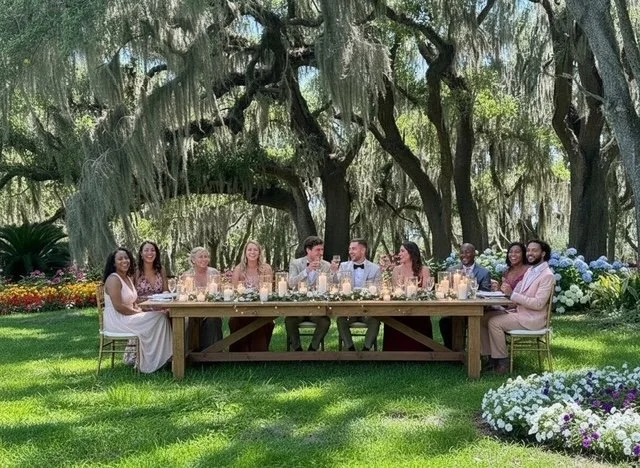 Group of ten people sitting at a long wooden table outdoors, under large trees with hanging moss, celebrating at a daytime event, with flowers and greenery around.