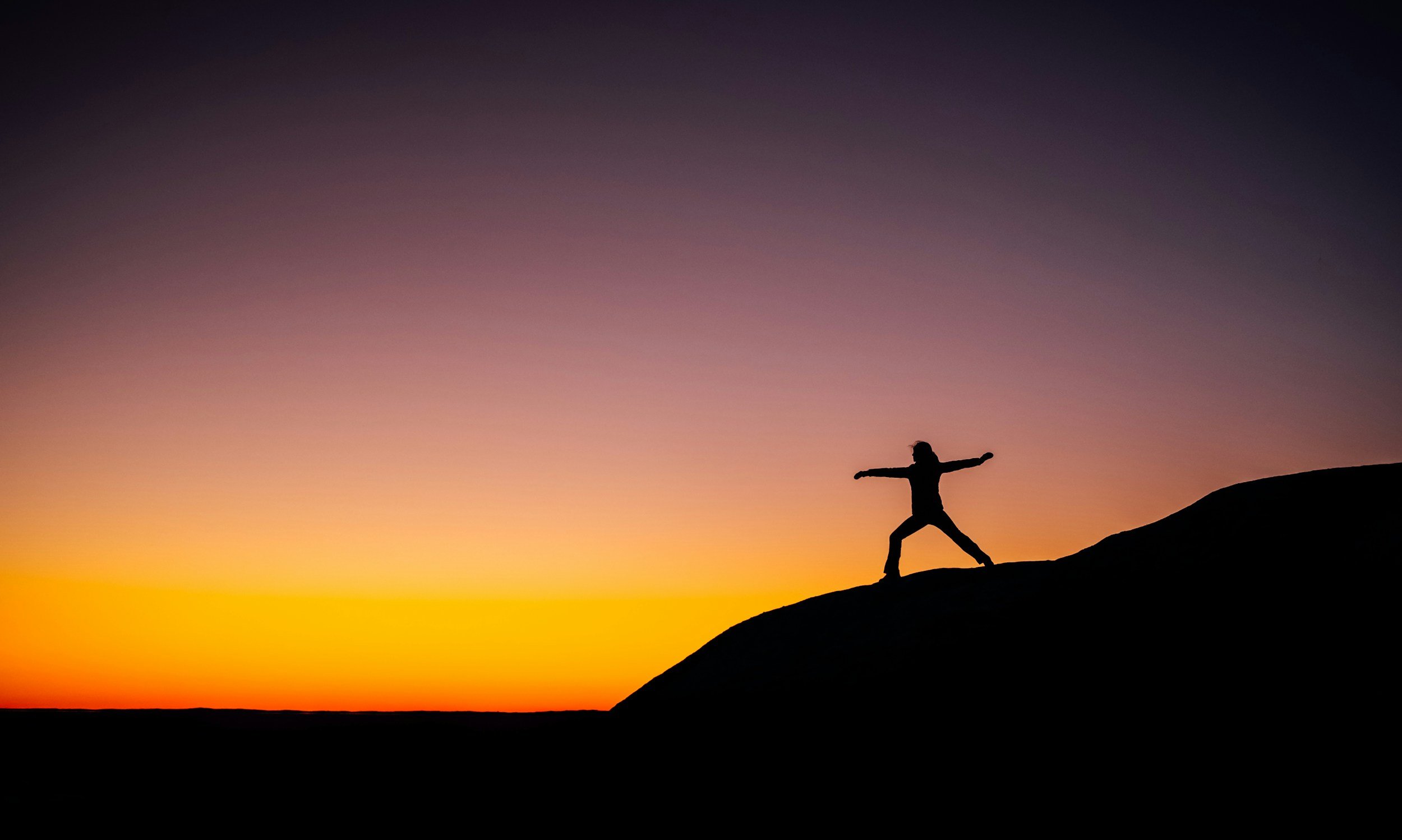 Person standing with arms outstretched on a hilltop during sunset, with a colorful sky of purple, pink, and orange hues.