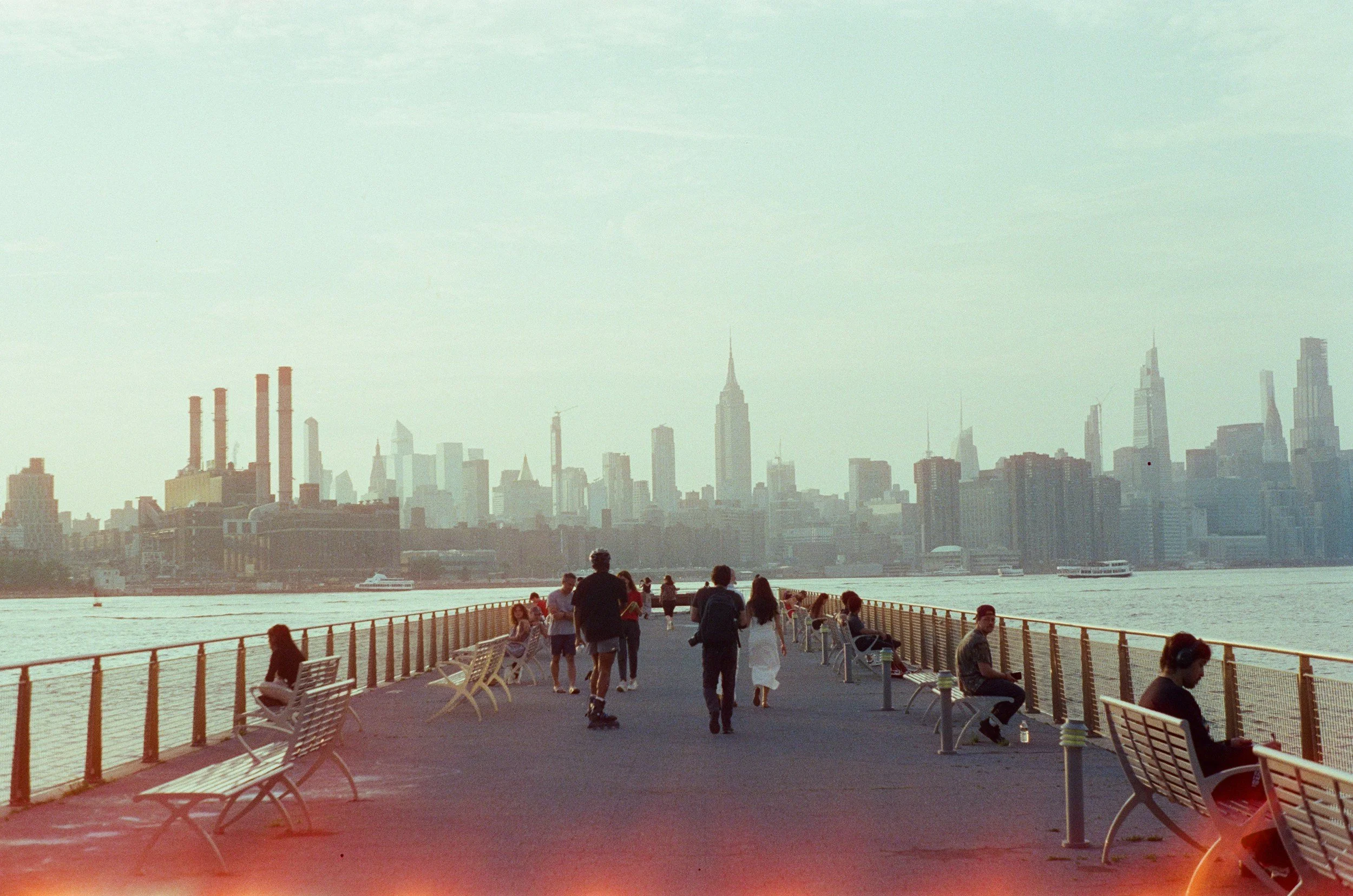 People walking and sitting on benches along a waterfront promenade with city skyline in the background, including skyscrapers and a river with boats.