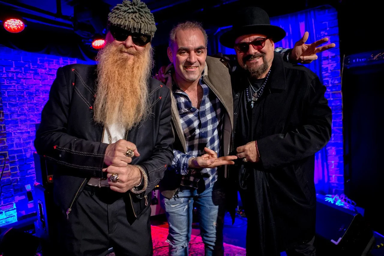 Three men posing on a stage with blue and purple lighting and a brick wall background. The man on the left has a long beard, sunglasses, a beanie, and a leather jacket. The man in the middle is smiling, wearing a checkered shirt and a jacket. The man on the right has a goatee, sunglasses, a hat, and multiple chains, making a peace sign with his hand.
