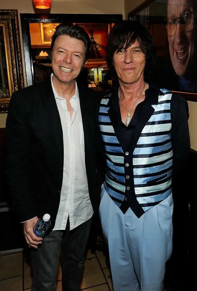 Two men posing for a photo in a restaurant or bar, smiling, with a framed mirror and wall decorations behind them.