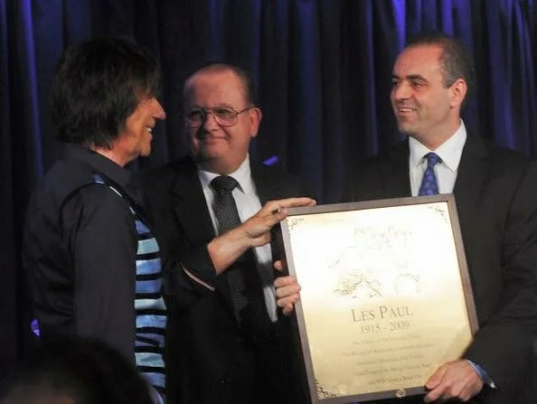 Three men in formal attire at a presentation, one holding a framed plaque.
