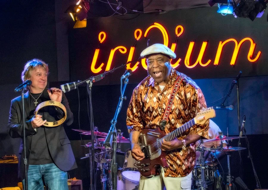 Musicians performing live on stage at the Troubadour, with a man playing guitar and singing, and a woman holding a tambourine. There is a neon sign in the background that reads 'Troubadour'.
