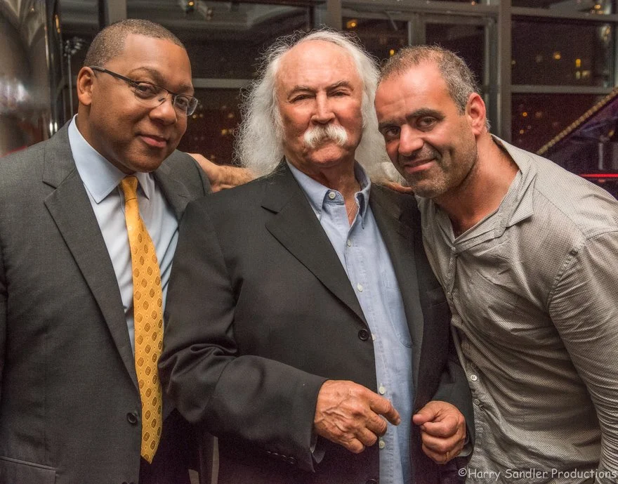 Three men posing together at an indoor event, with a dark background and warm lighting.