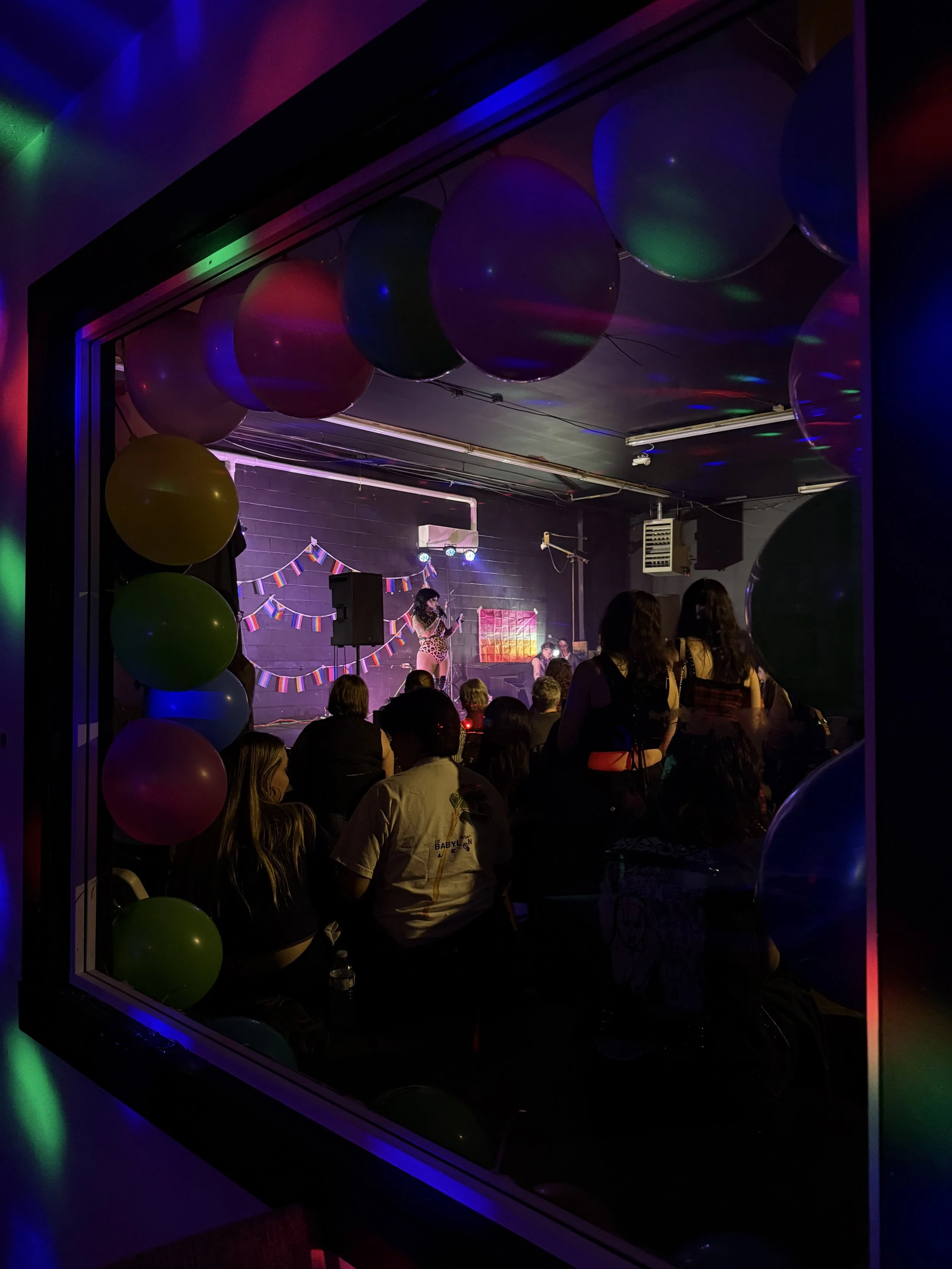 People gathered in a dark indoor venue decorated for a celebration, with balloons and a colorful banner, while a performer on stage entertains the audience.