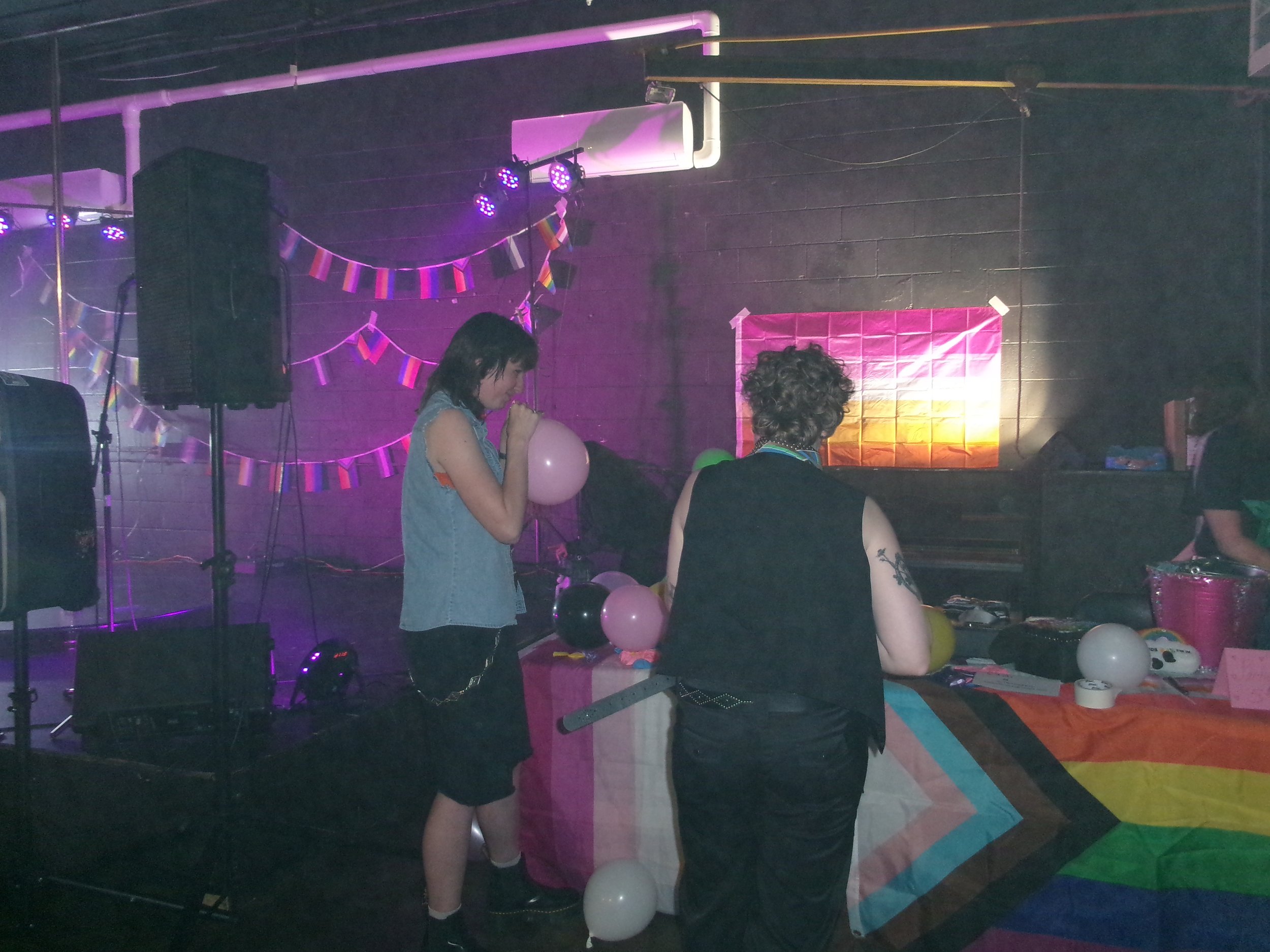A party scene with two women, one holding balloons, standing near a table with balloons and decorations, under purple lighting, with a black wall, speakers, and rainbow flags in the background.