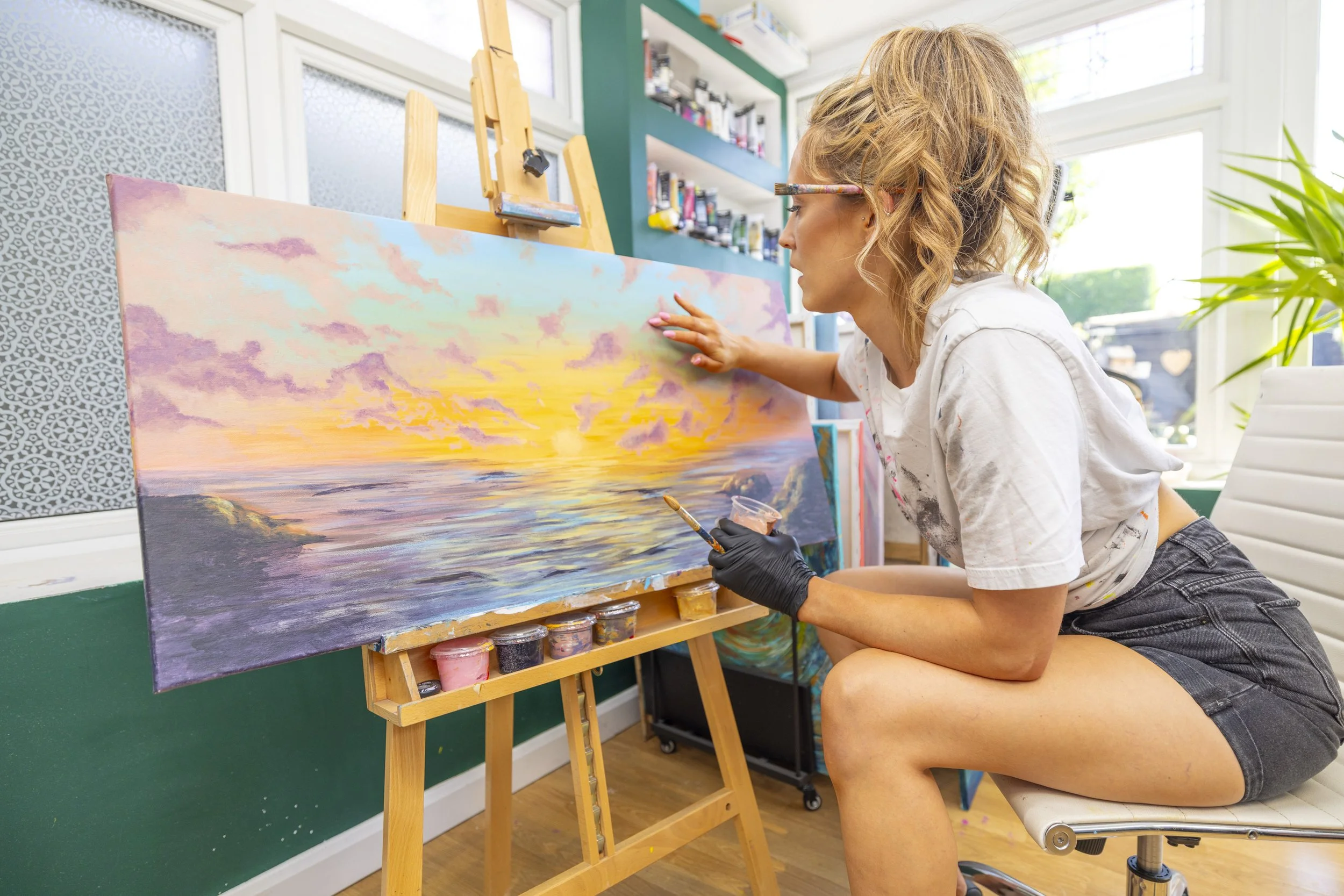 A woman painting a sunset scene on a large canvas in an art studio, wearing glasses and a white t-shirt, sitting on a rolling stool.