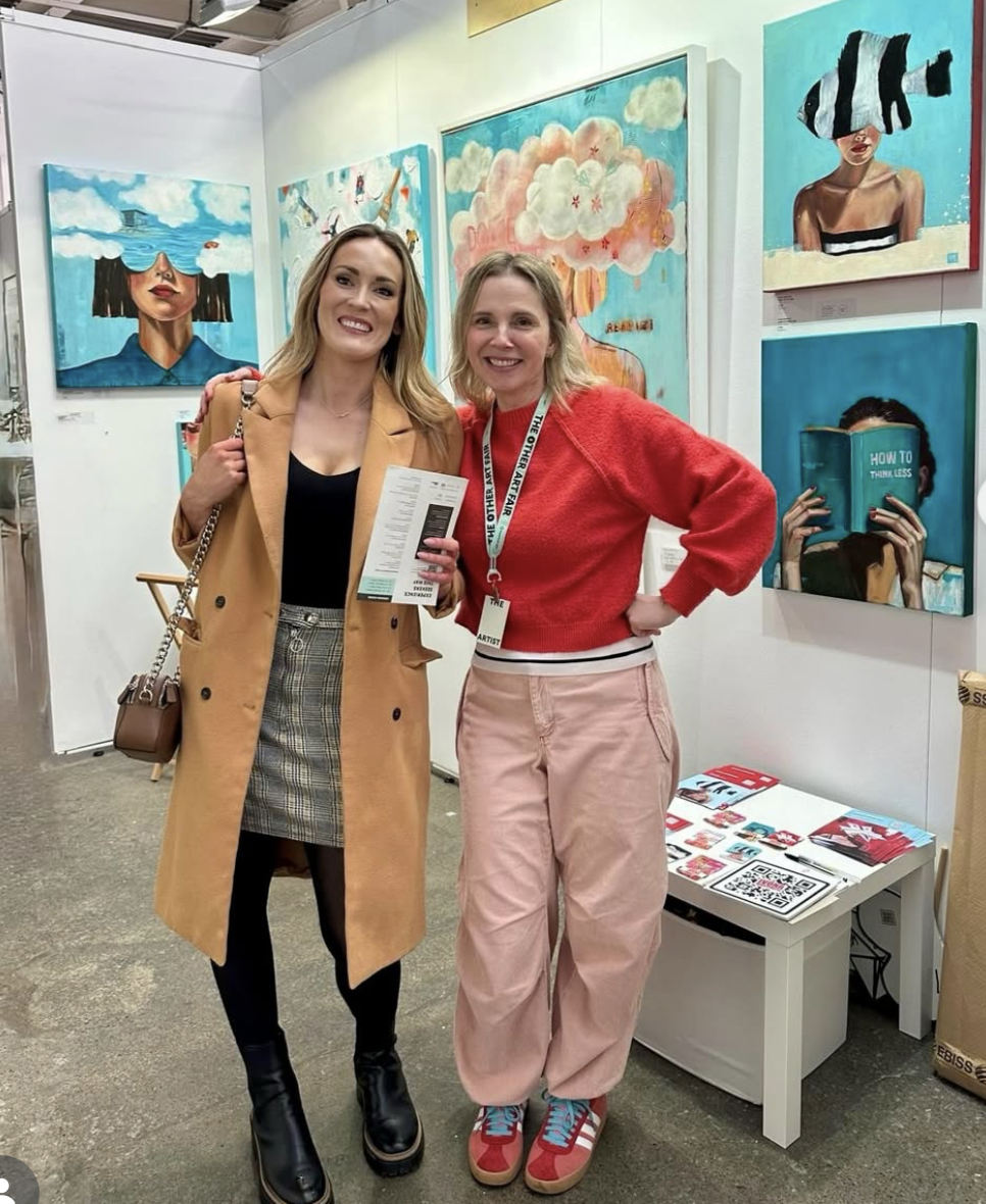 Two women standing in front of colorful artwork at an art exhibit, smiling and posing for the photo.