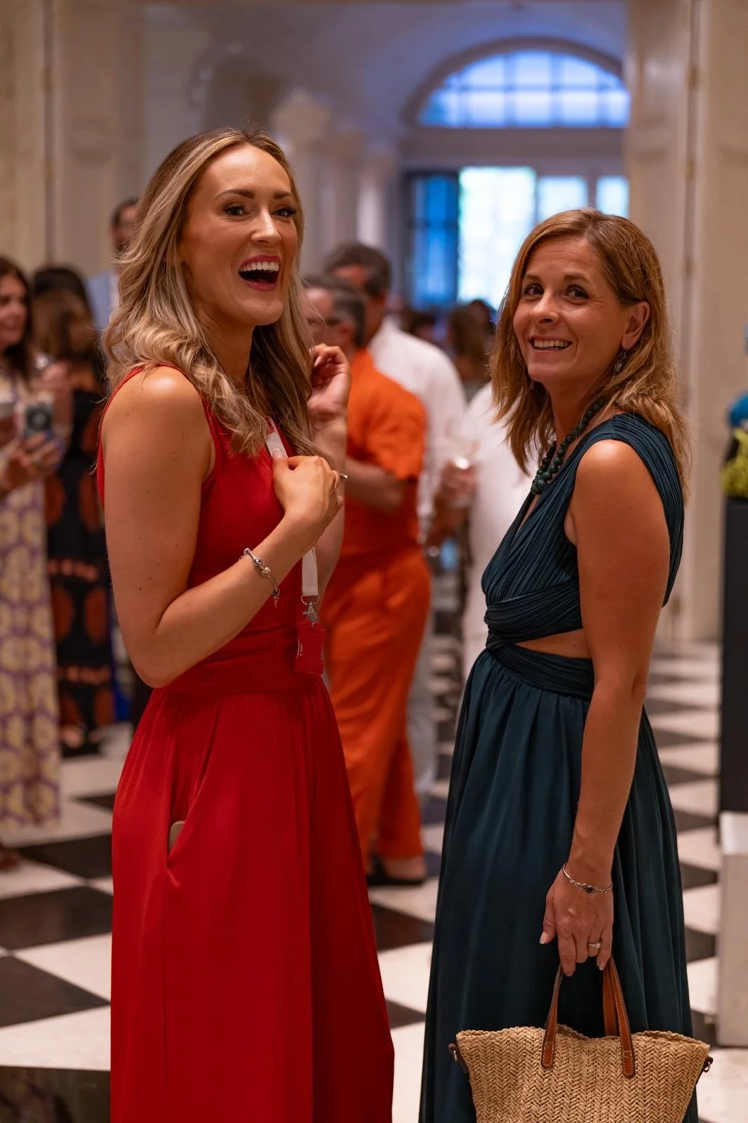 Two women smiling at each other at a social event, with people in the background. One woman is wearing a red dress and the other a dark blue dress, both in an elegant setting.