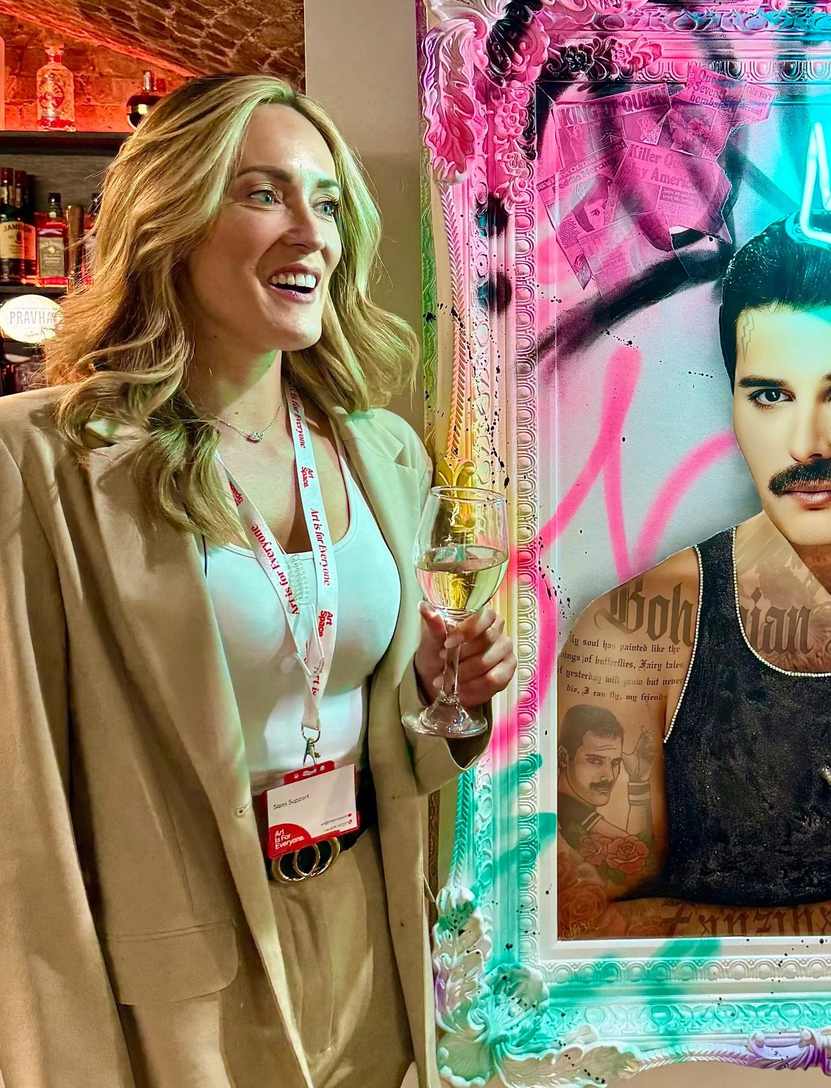 A woman with blonde hair, wearing a beige blazer and white top, holding a glass of white wine, standing next to a colorful, neon-lit portrait with various images and text, in an indoor setting with shelves of liquor bottles in the background.