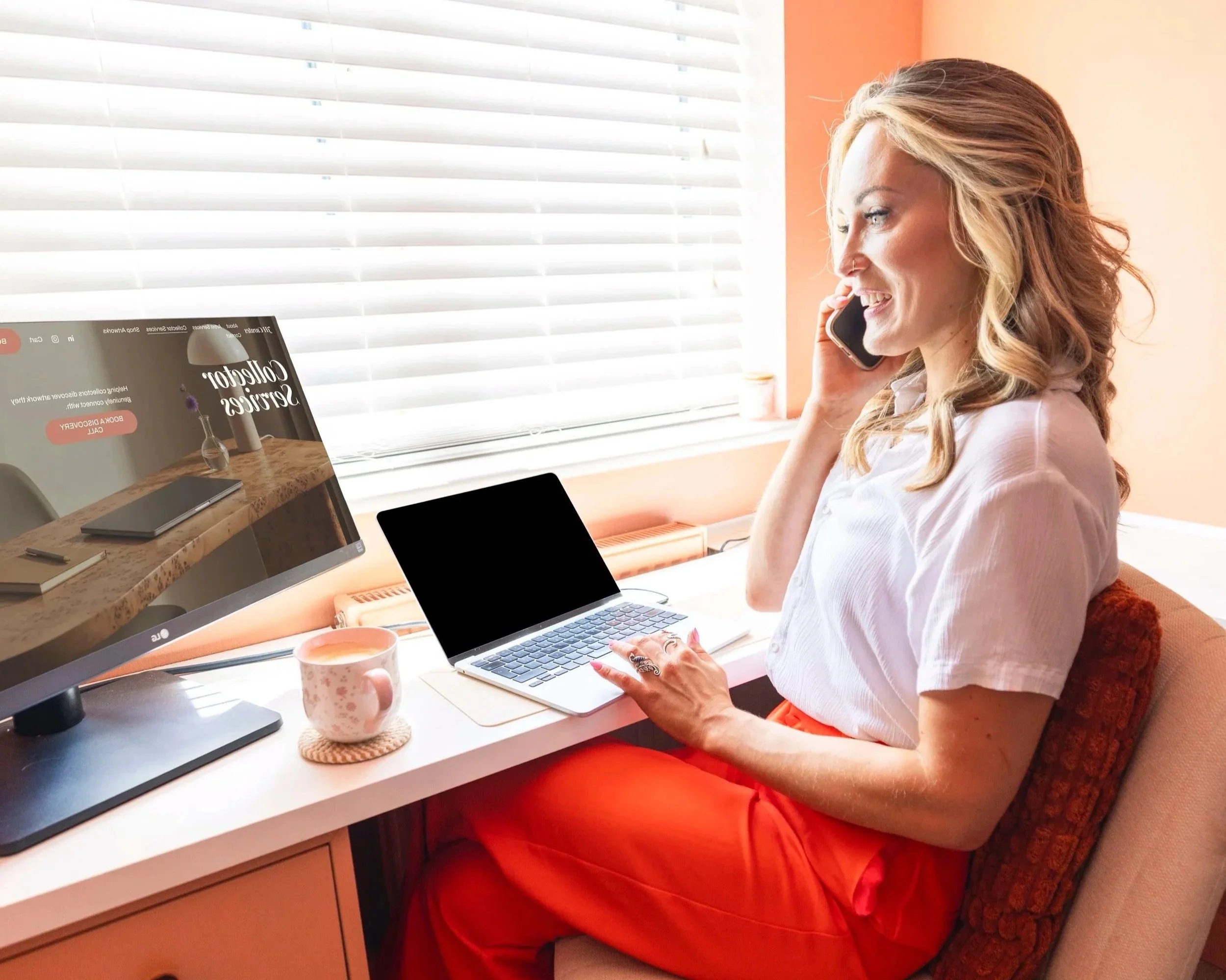 A woman with long blonde hair, wearing a white shirt and red pants, sitting at a desk with a laptop, talking on the phone, with a notebook, a mug, and a monitor on the desk.