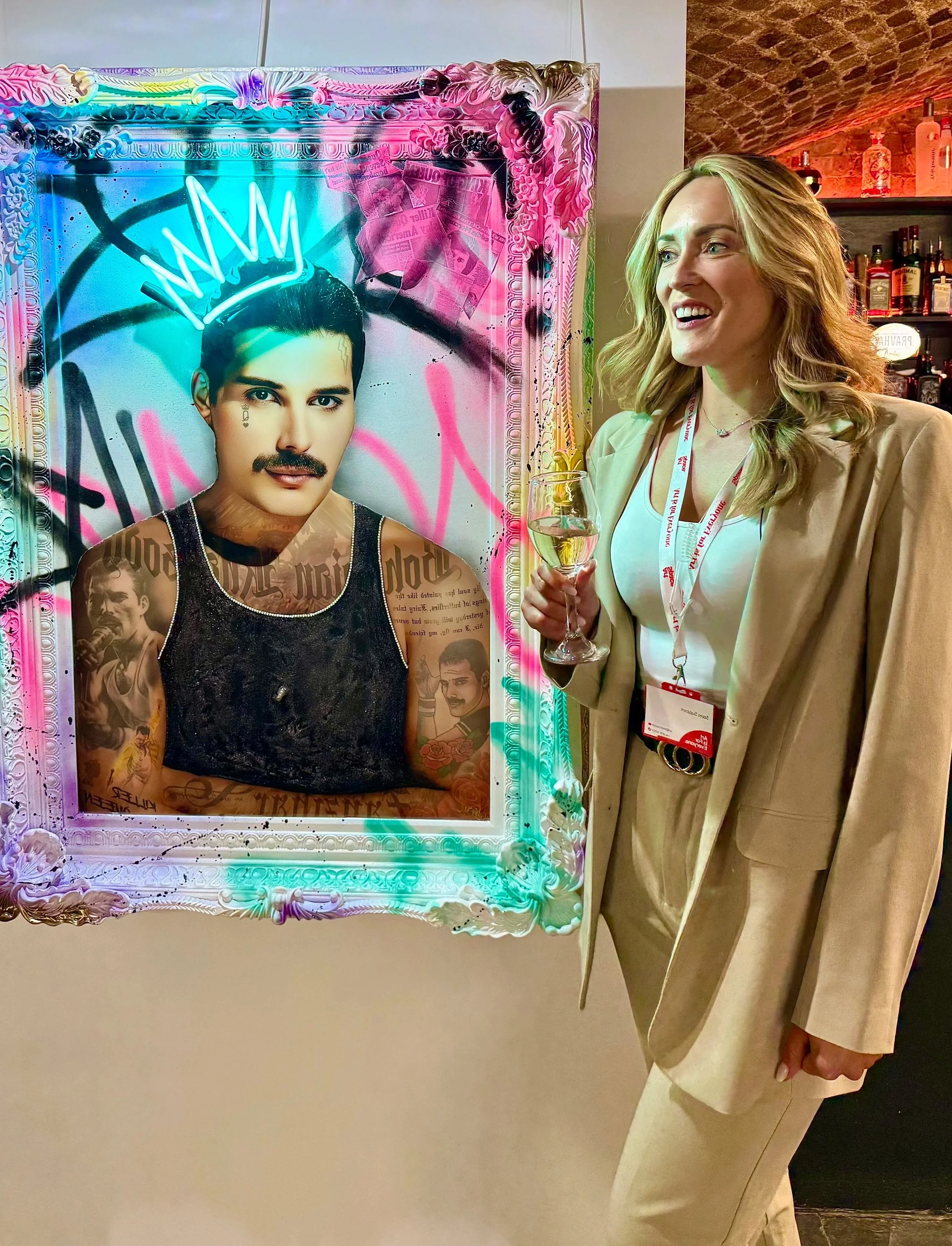 A woman holding a glass of white wine standing next to a colorful, graffiti-style artwork of a person's face with a crown-shaped glow above their head inside a bar or restaurant.