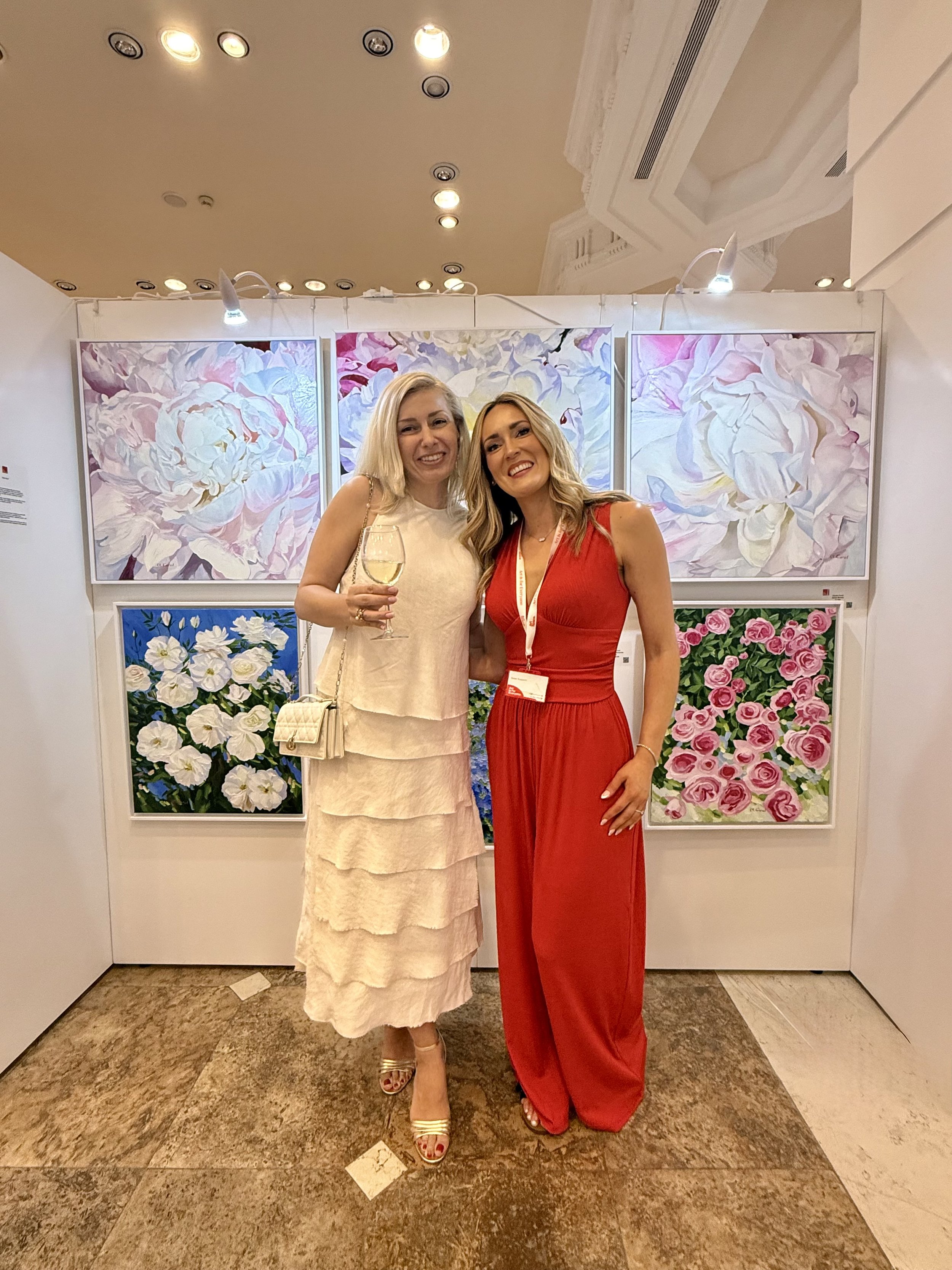 Two women standing together, smiling at the camera, in front of floral artwork at an art gallery, with one woman holding a glass of white wine, both dressed elegantly.