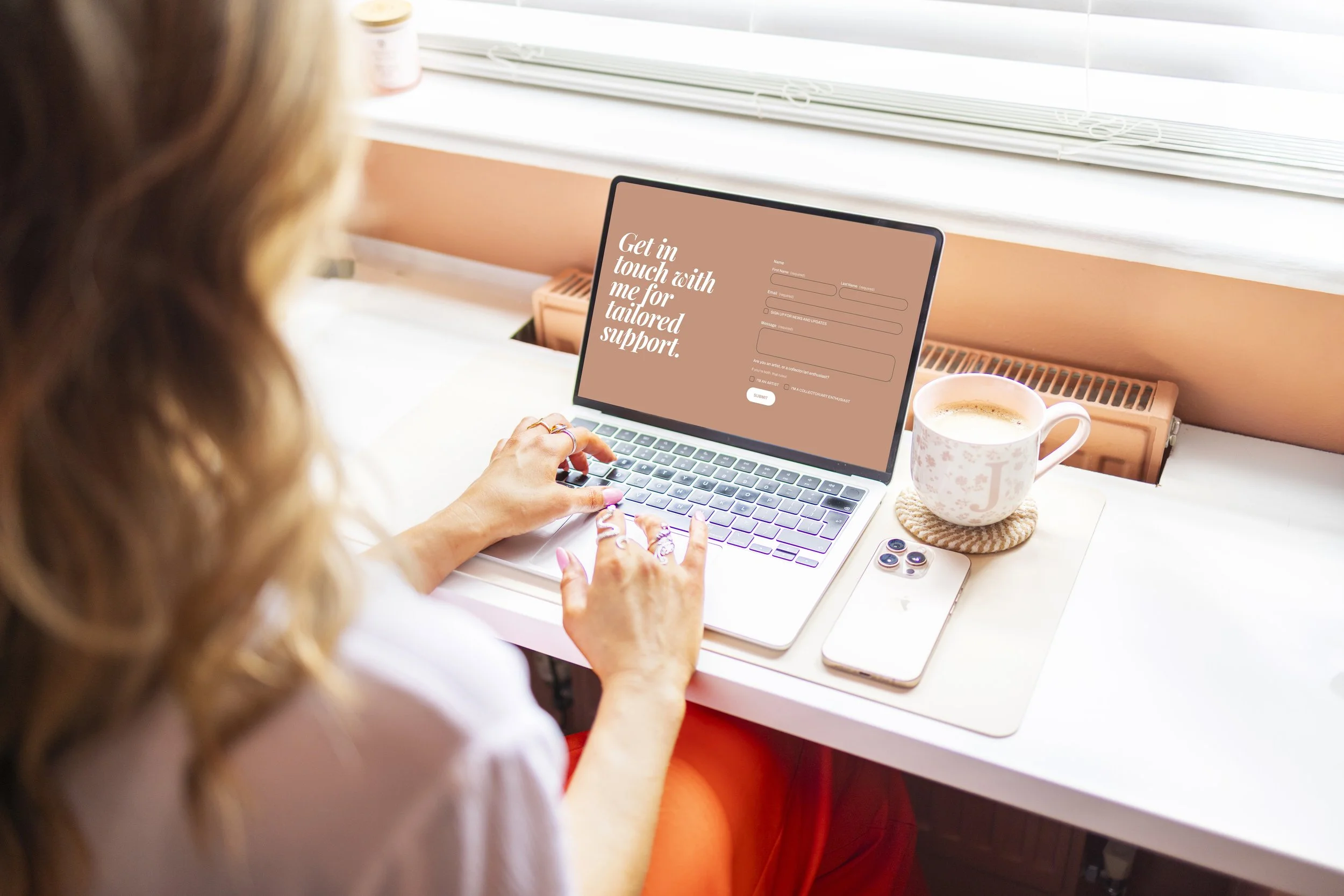A person with long reddish hair using a laptop with a pinkish screen that displays a form for tailored support, sitting at a white desk with a mug and small items, near a window with white blinds.