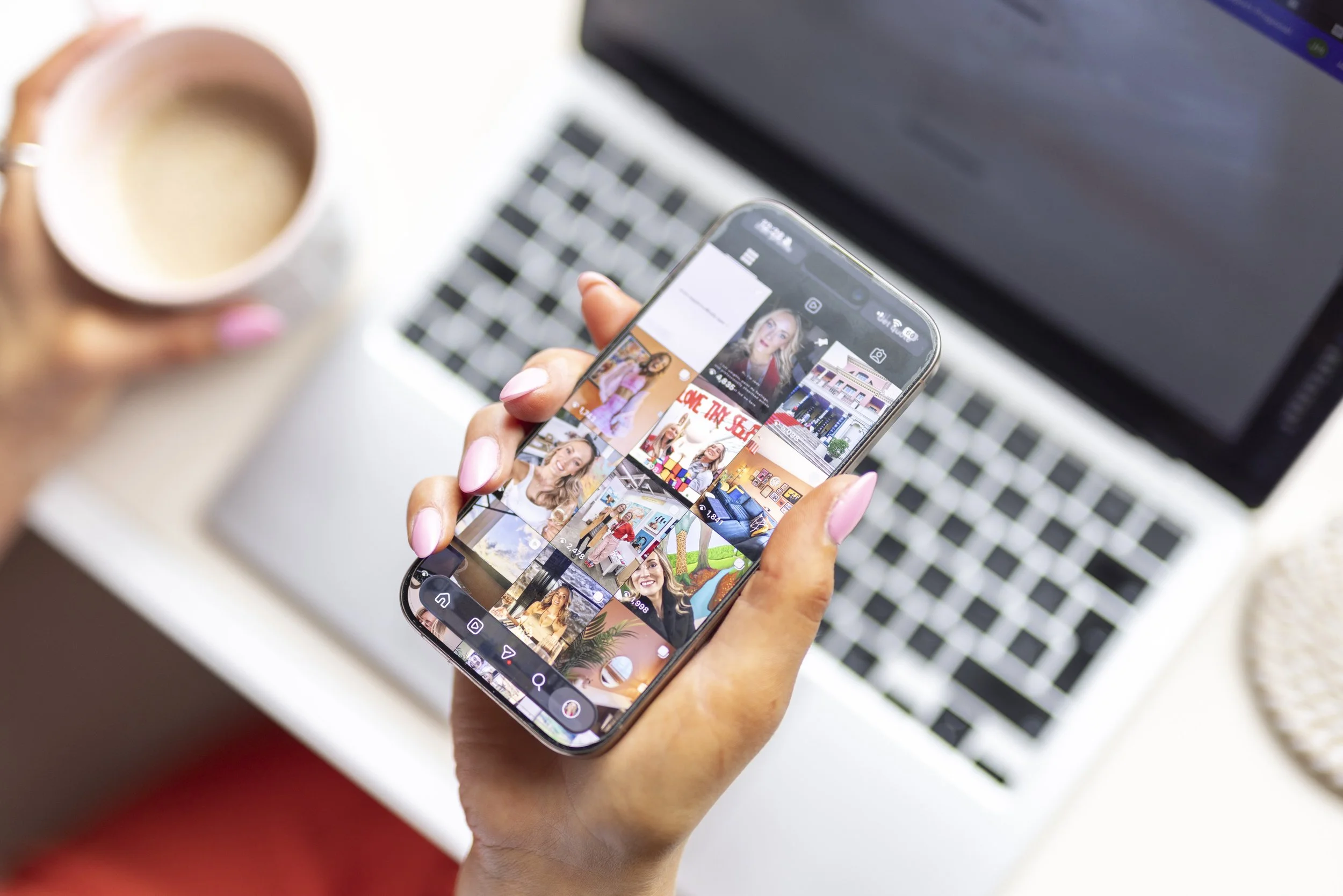 Close-up of a person with pink painted nails holding a smartphone displaying a photo gallery, with a laptop and a mug of coffee in the background.