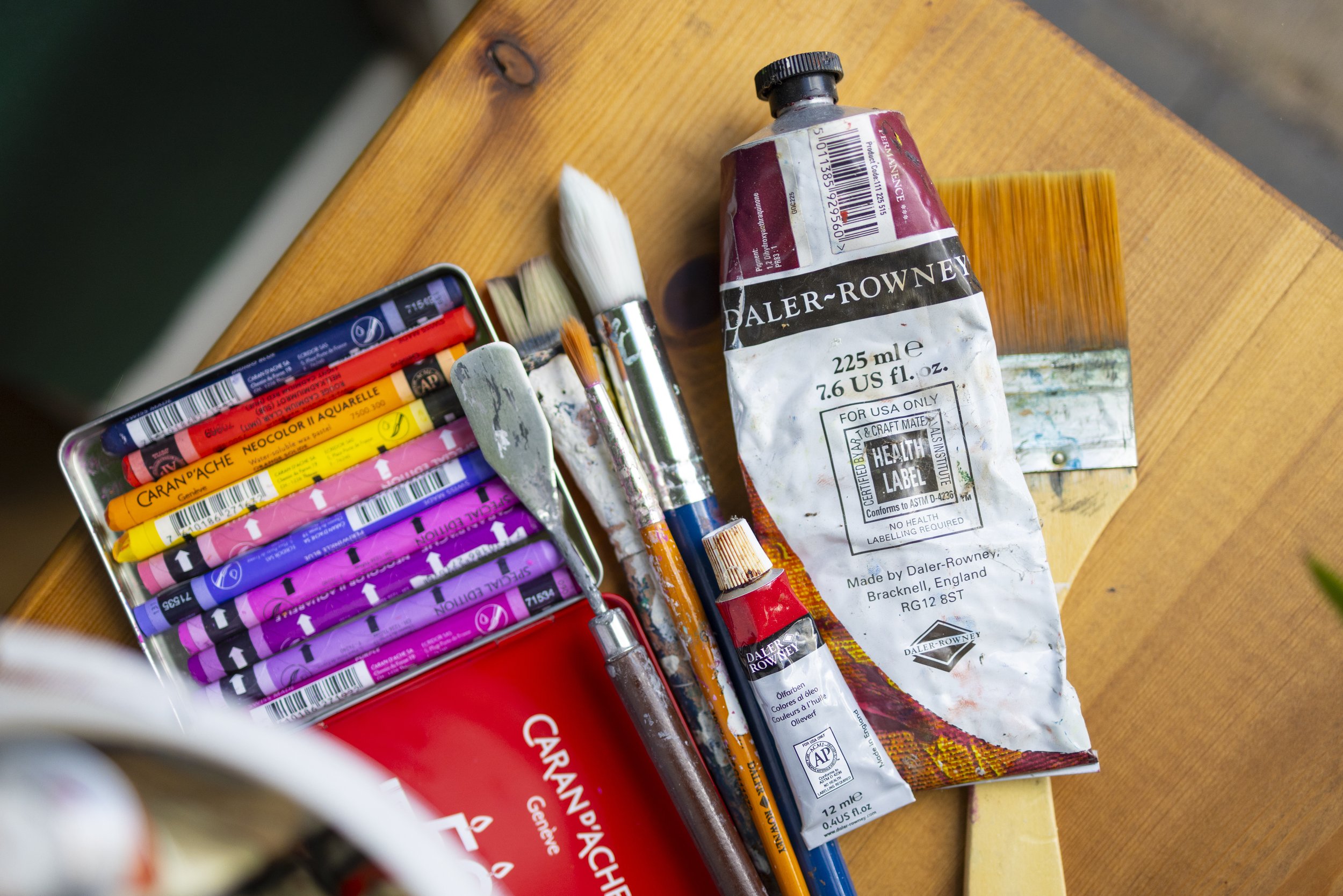 A wooden table with a metal box of colorful oil pastels, paintbrushes, and a crumpled tube of paint on it.