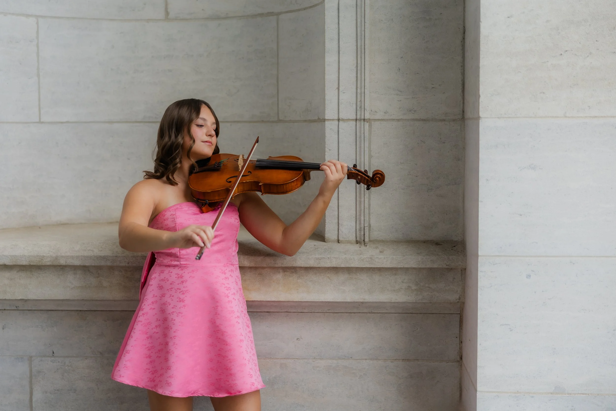 A young girl in a pink dress playing a viola while standing against a beige stone wall.