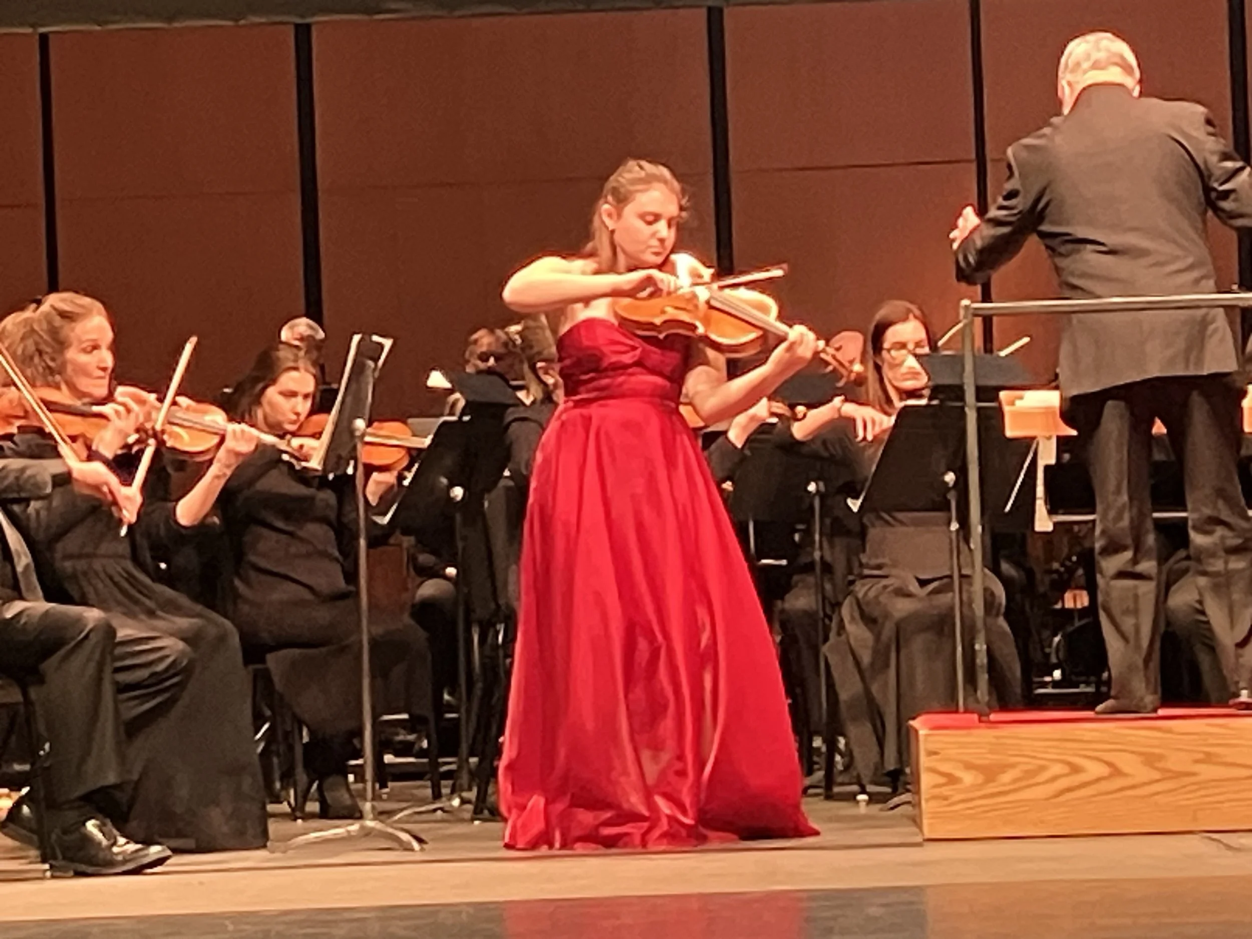 A woman in a red dress playing the viola on stage during an orchestral performance, with conductor and other musicians in the background. Des Moines Symphony