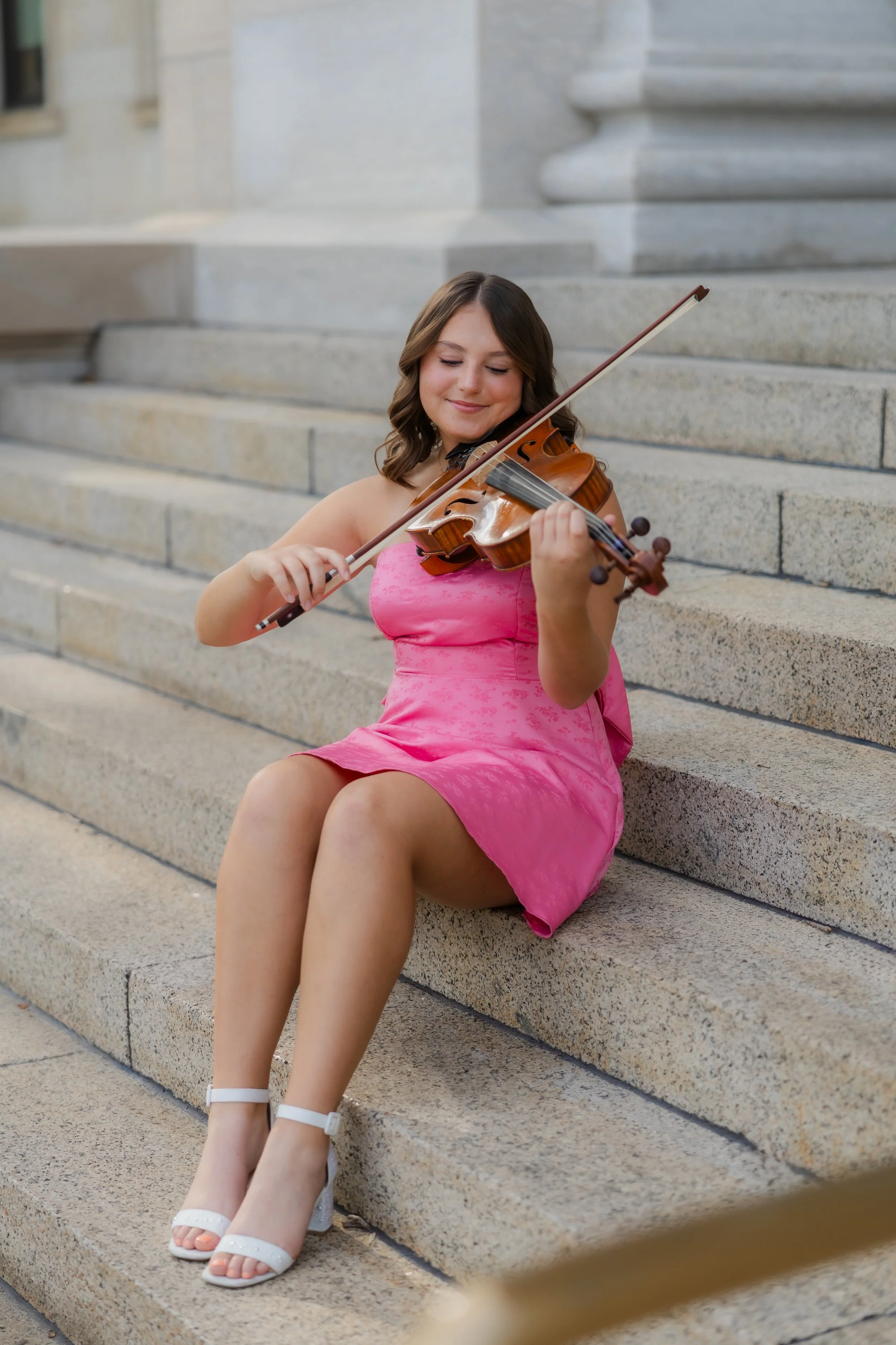 Young woman with brown hair in a pink dress playing the violin on stone steps outside a building.