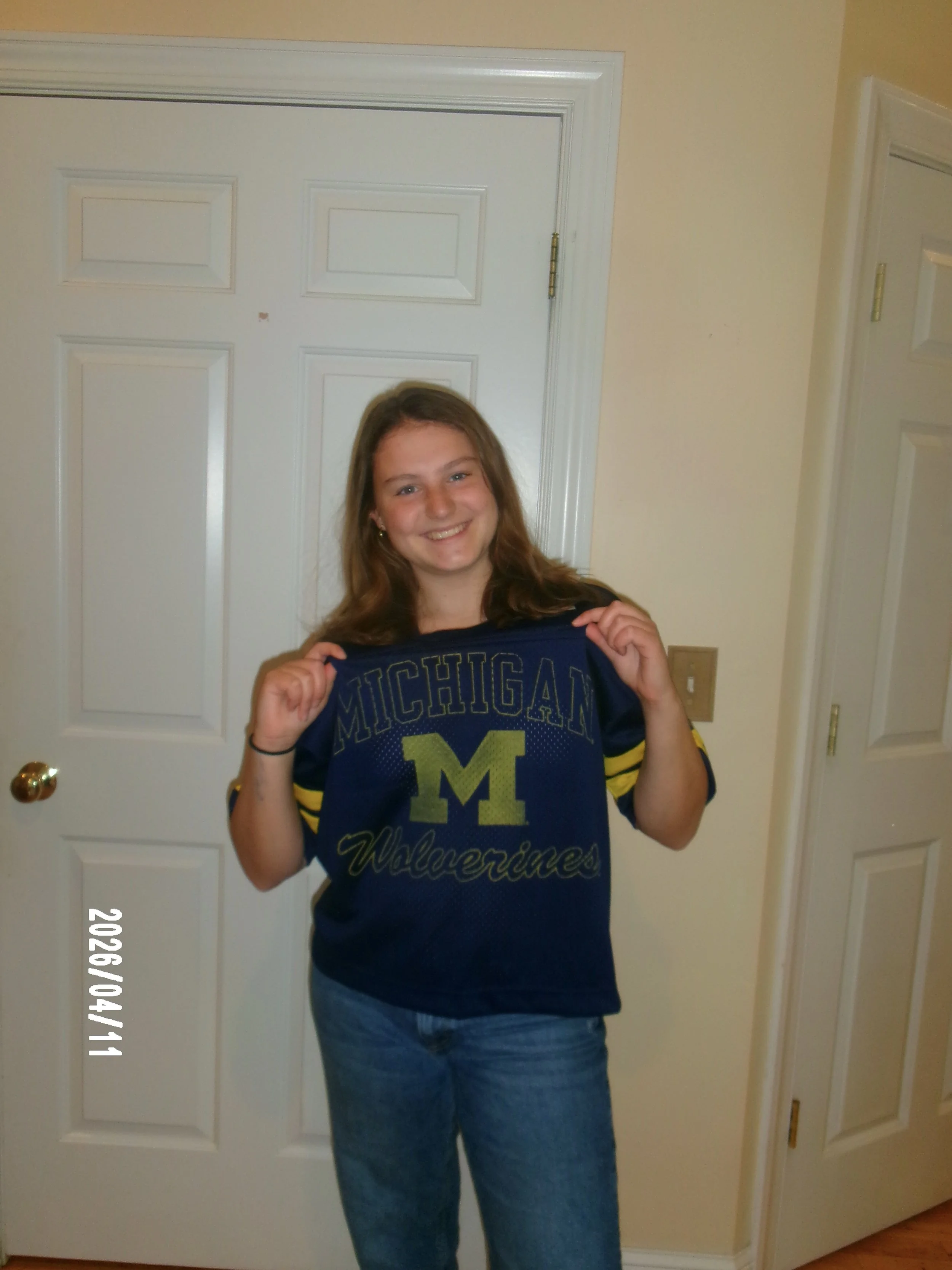 A young woman with brown hair smiling, holding a Michigan Wolverines T-shirt in front of her chest, standing in front of a white door in a room with beige walls.