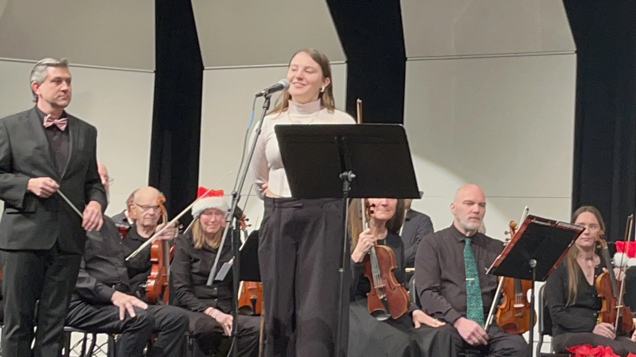 A woman talking into a microphone on stage with an orchestra group behind her, including musicians holding violins and a conductor standing to the left.