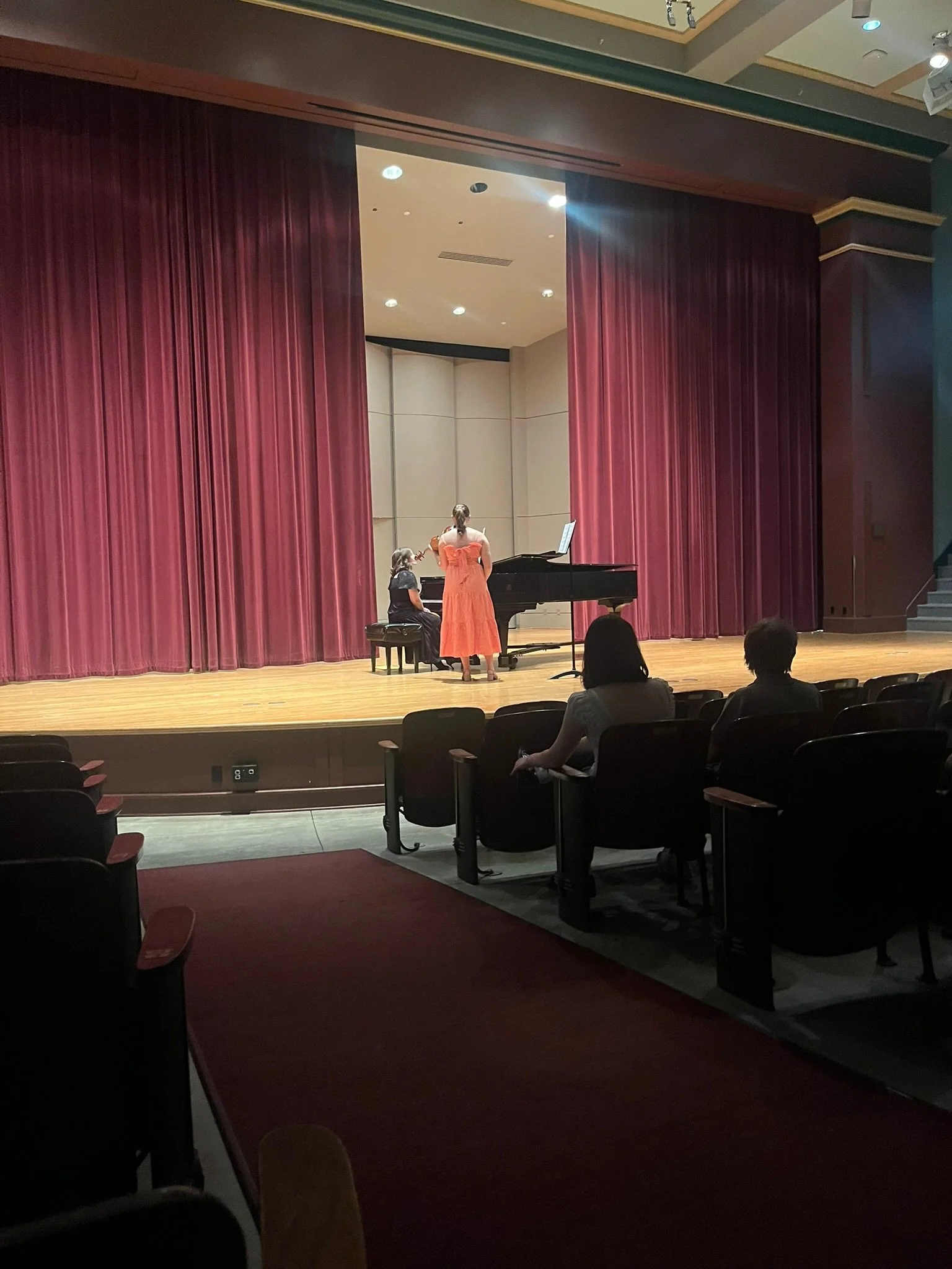 Two women are performing a musical piece on a stage with a grand piano and red curtains. One woman is seated at the piano, while the other woman is standing and playing a violin.