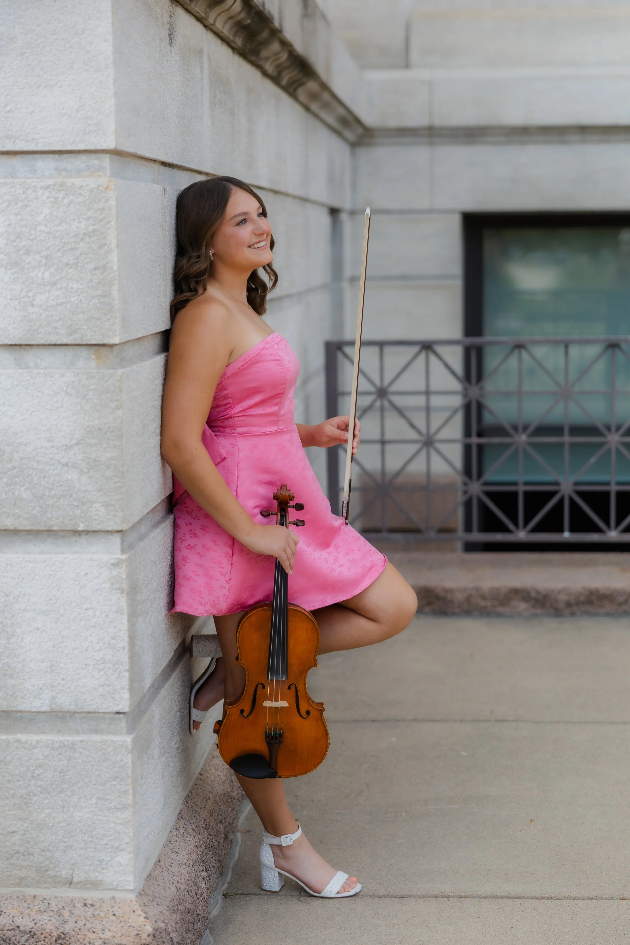 A young woman in a pink dress, white high heels, and jewelry, leaning against a light-colored stone wall, holding a viola in one hand and a bow in the other, smiling while looking upwards.