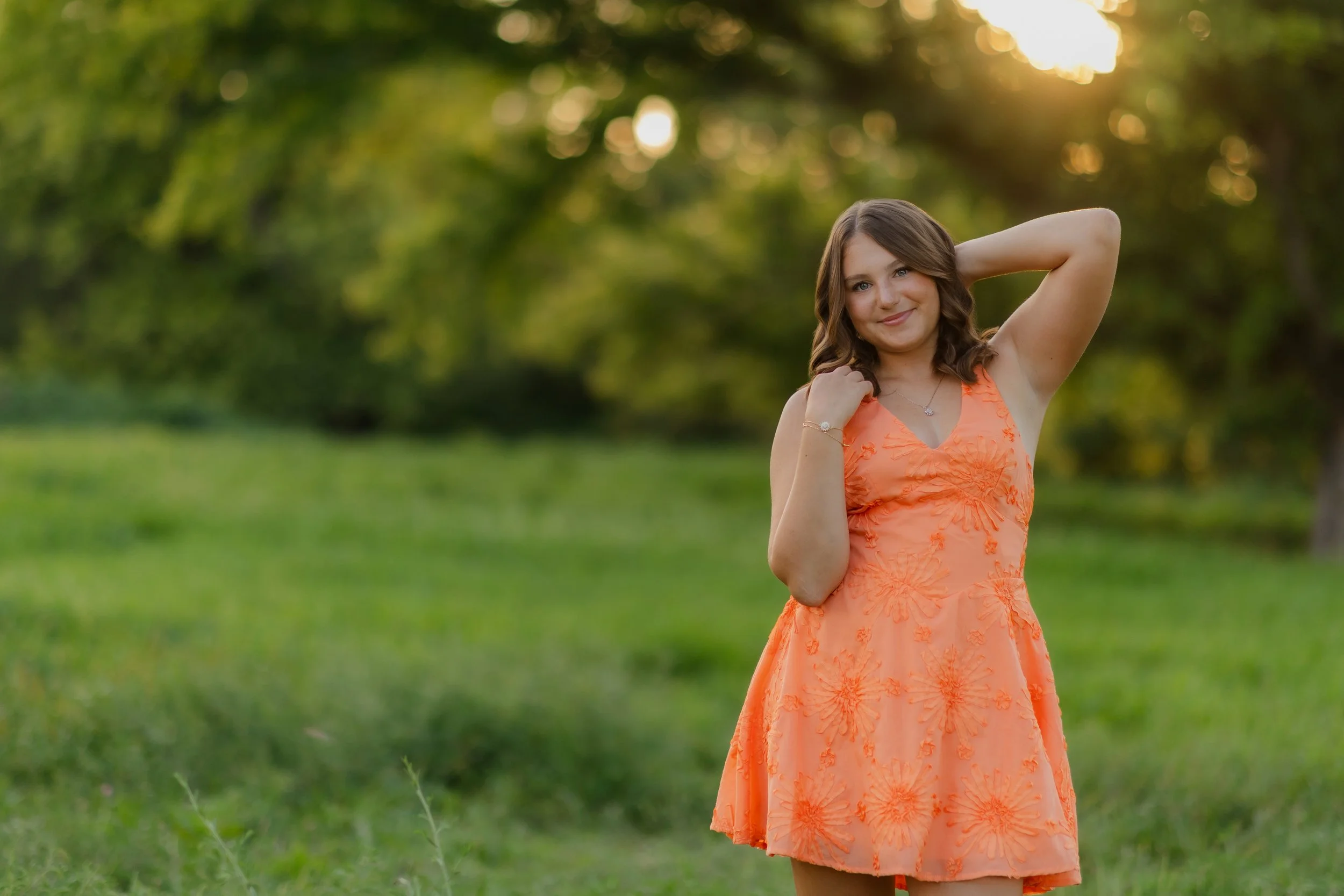 A young woman with brown hair smiling outdoors in a green park during sunset, wearing an orange dress with floral embroidery.