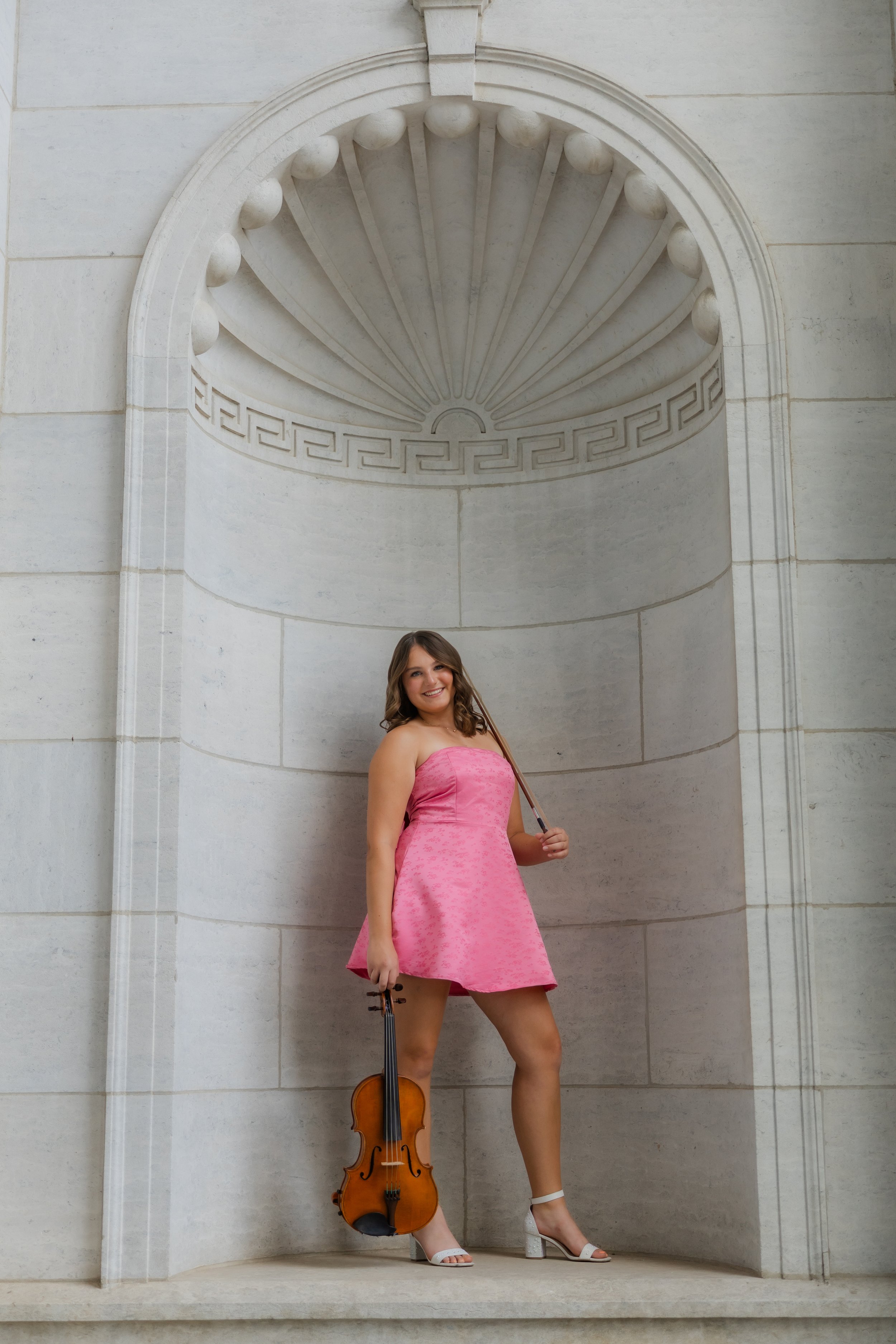 A young woman in a pink strapless dress is standing in an architectural alcove with a large shell-shaped ornament above her. She is holding a violin at her side and wearing white high-heeled shoes, with a smile on her face.
