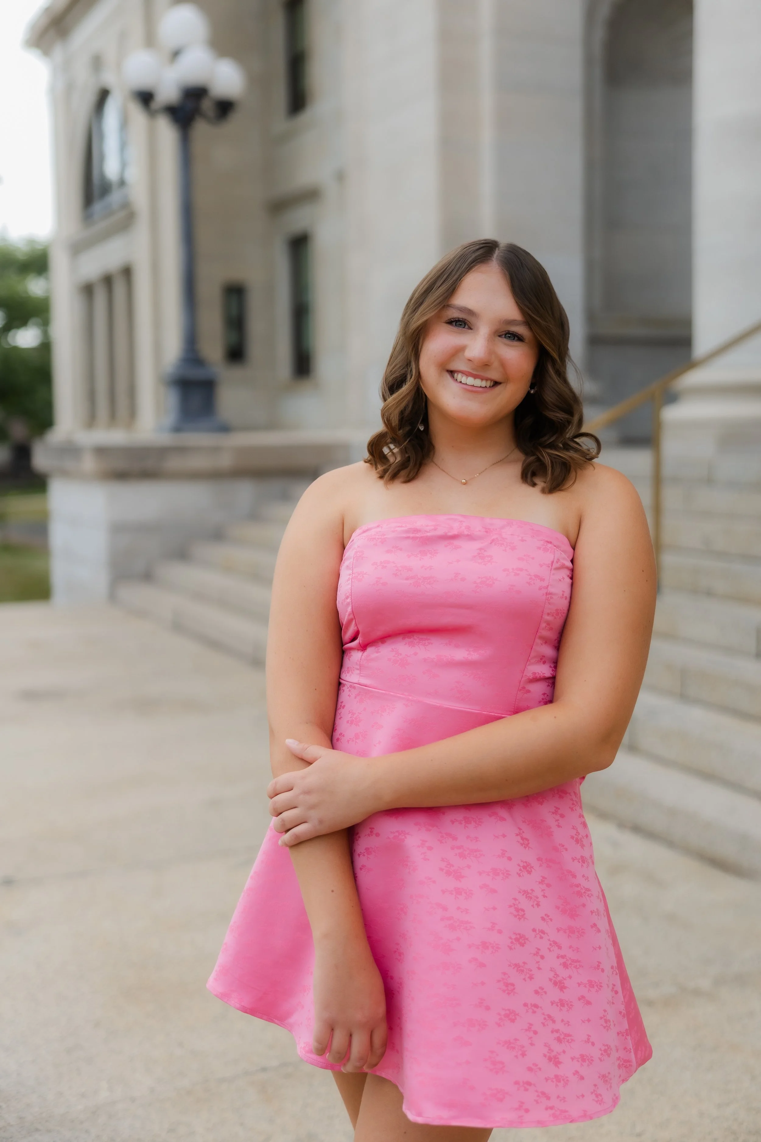 A young woman in a pink dress standing outside in front of a building with stairs and large windows, smiling at the camera.