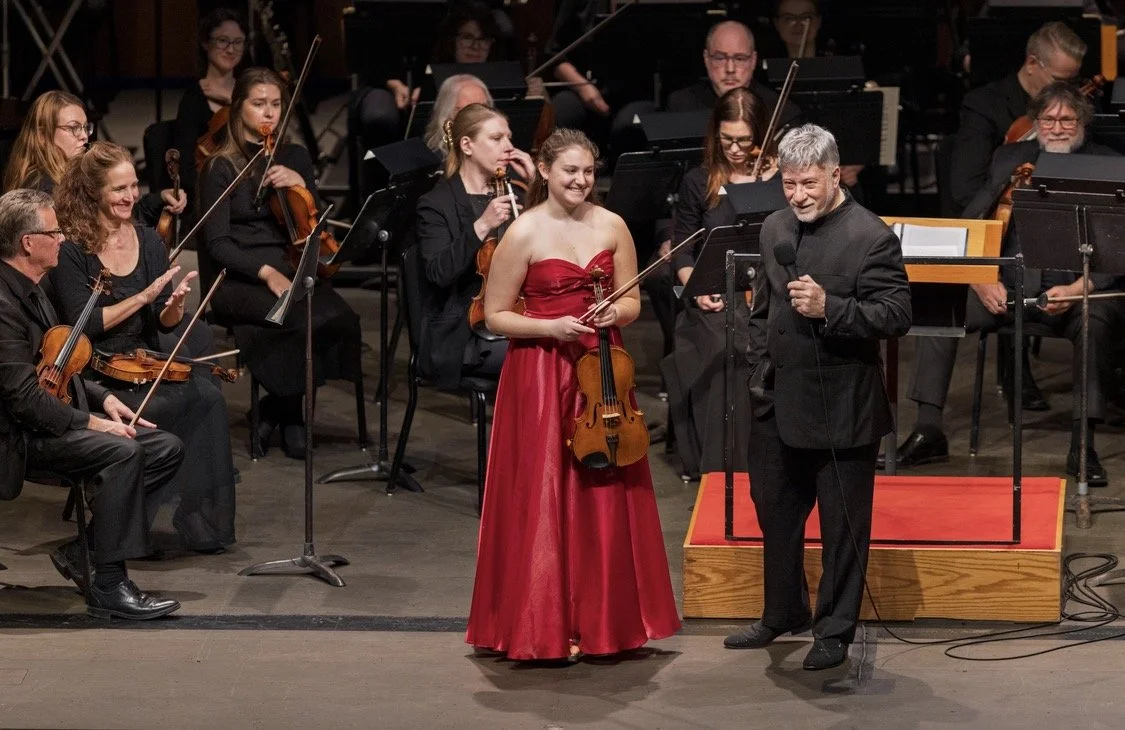 Orchestra on stage with a woman in a red dress holding a violin and a man in black holding a microphone, surrounded by musicians with string instruments.