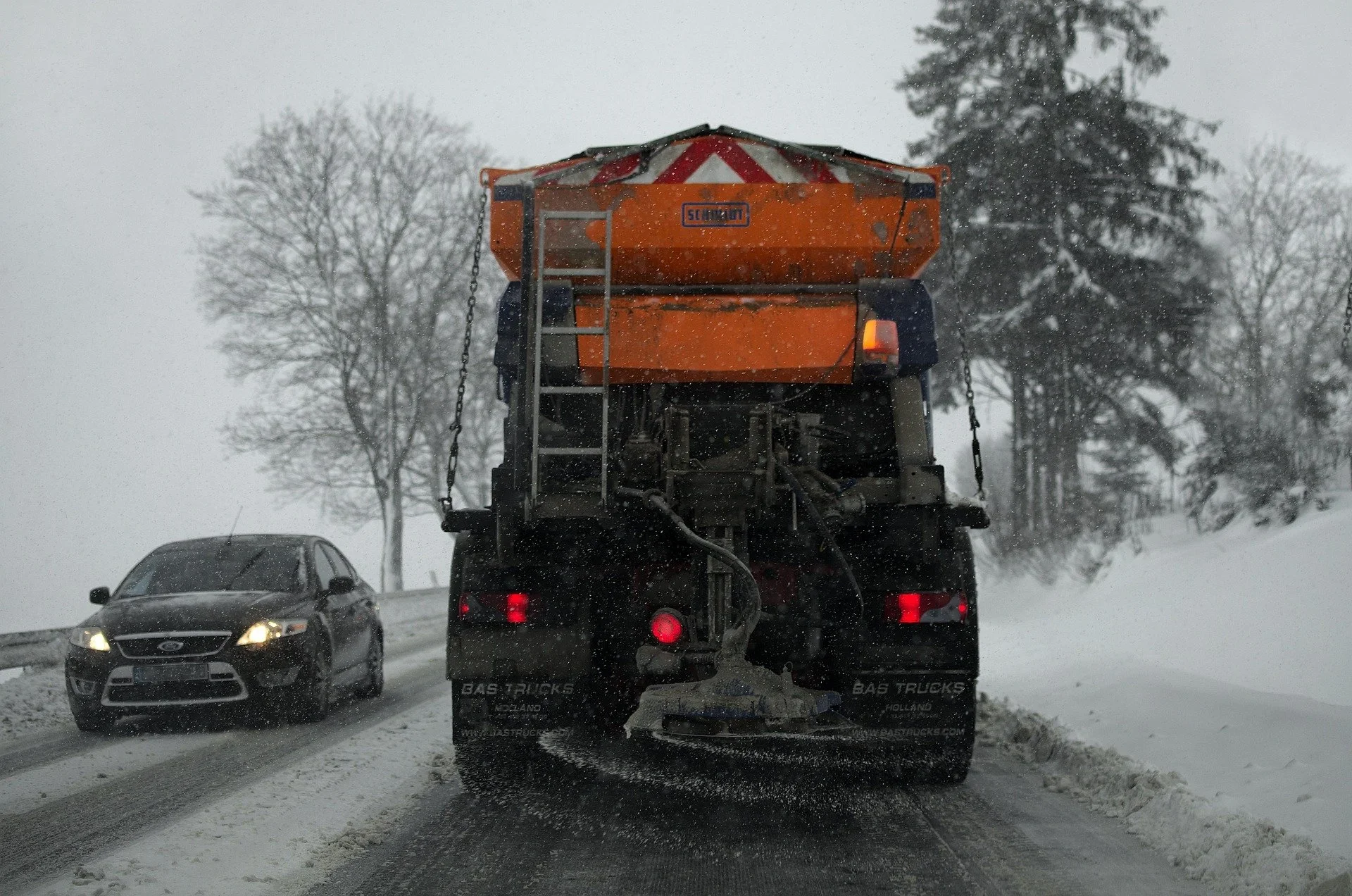 Snowy road with vehicles, including a black sedan and a snow plow truck, during snowfall with bare trees in the background.
