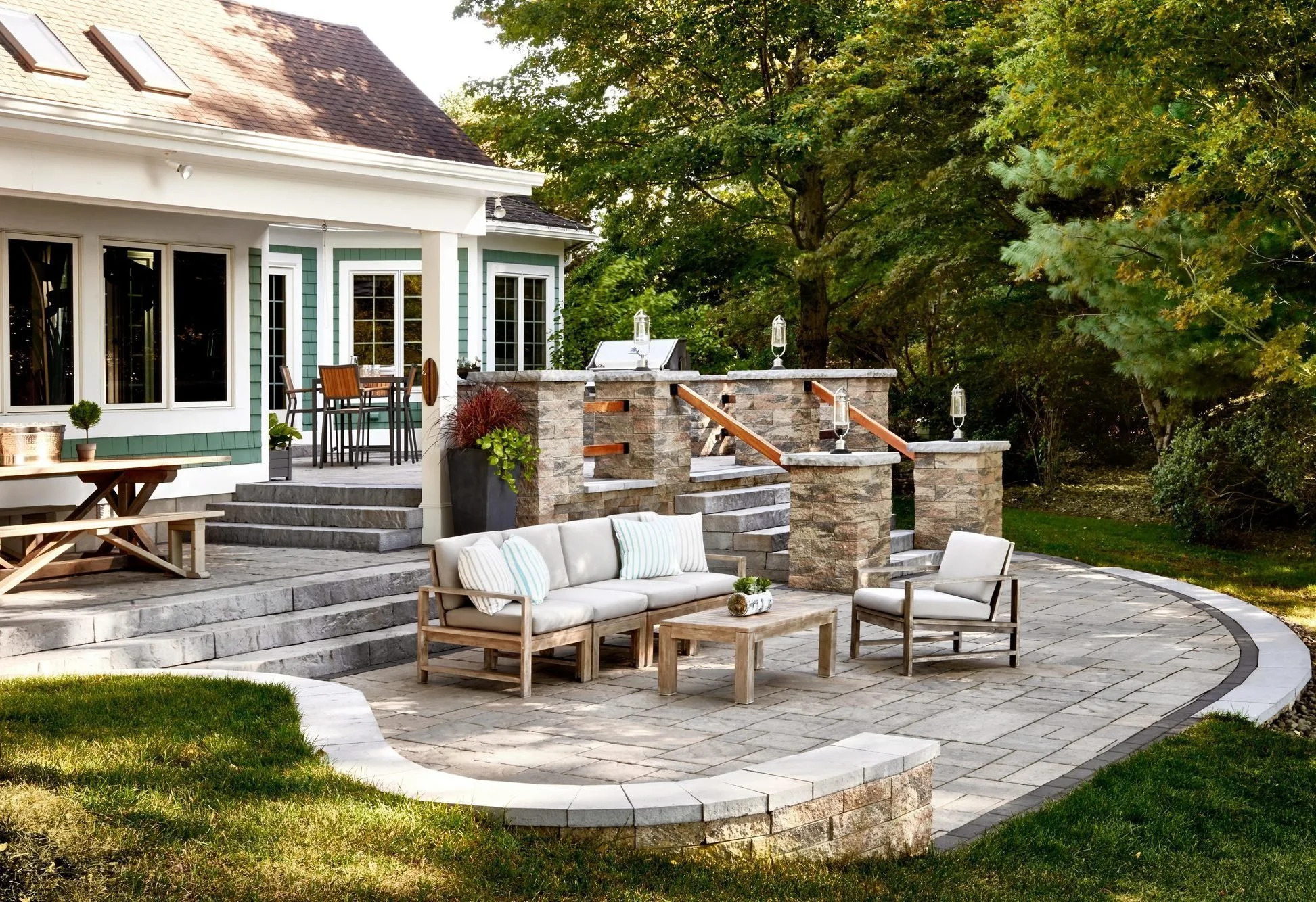 A spacious backyard patio with stone steps and a curved retaining wall, furnished with a cushioned outdoor sofa, chairs, and a coffee table, surrounded by green trees and a house with sliding glass doors and a grill.