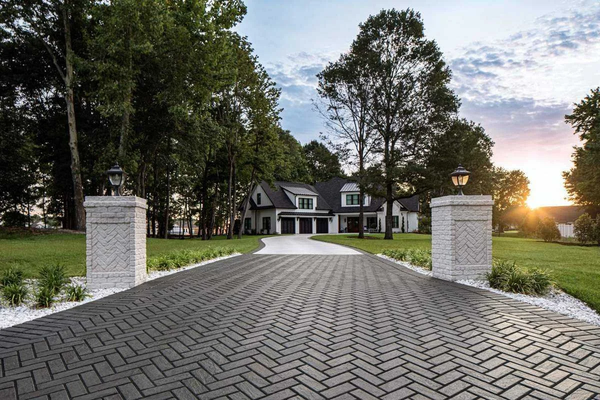 A paved driveway with brick pattern leading to a large, modern house surrounded by trees and green lawn, with decorative white brick pillars and lanterns at the entrance at sunset.